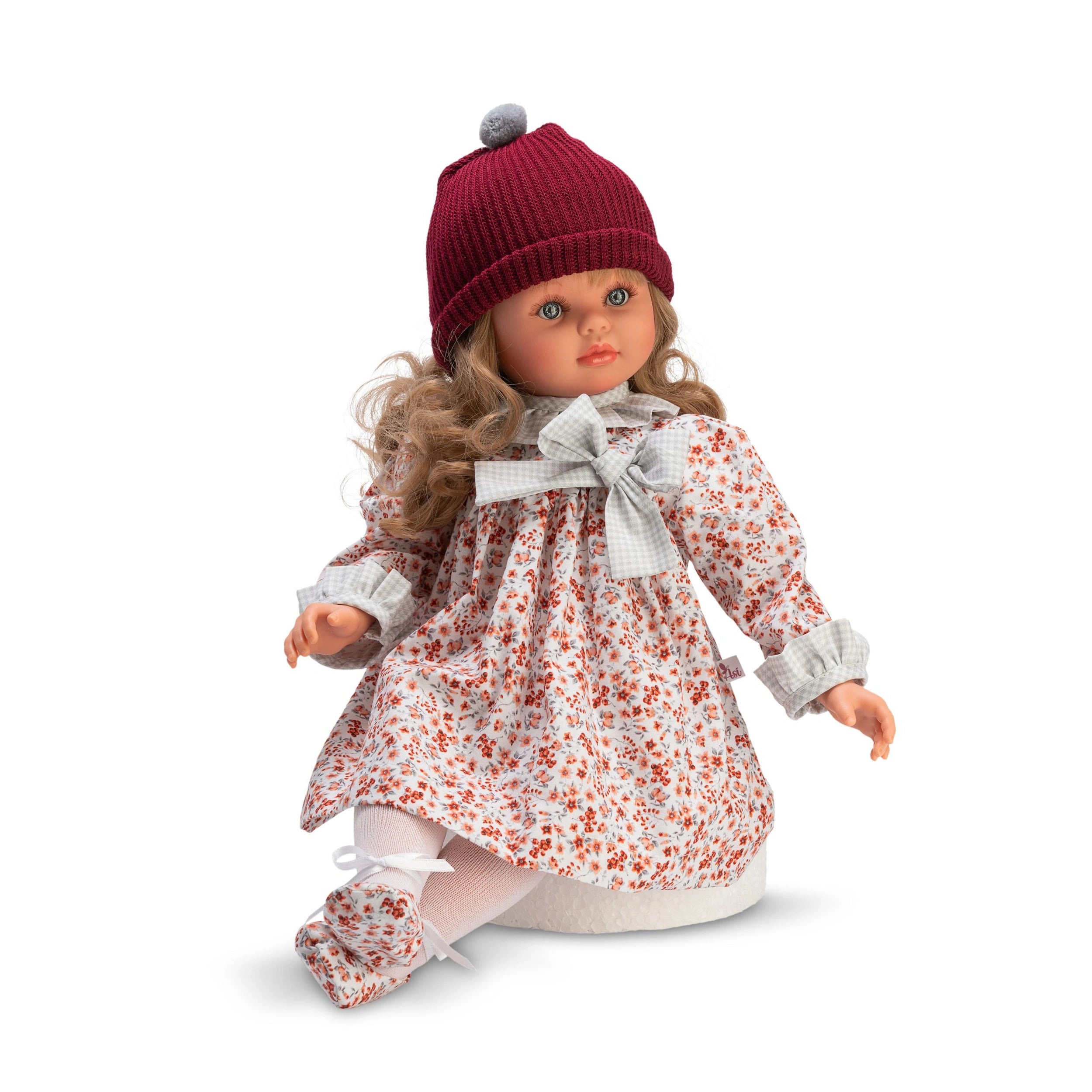 Pepa doll in floral dress and maroon hat, with long curly hair and expressive eyes, seated with one knee up.