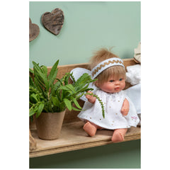 ASI Bomboncín Guardian Angel doll with blonde hair, white dress, and wings, sitting on a wooden shelf beside a potted plant.