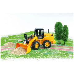 Yellow Bruder FR 130 articulated road loader with moving bucket on green diorama near trees and gravel piles.