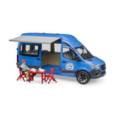 Blue Bruder MB Sprinter camper toy with open doors, retractable awning, and seated driver figure at a red table outdoors.