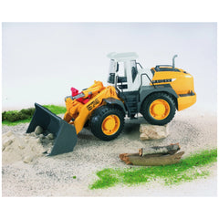 Bruder Liebherr L574 road loader toy with yellow body and tilting shovel, on sand with rocks and greenery.