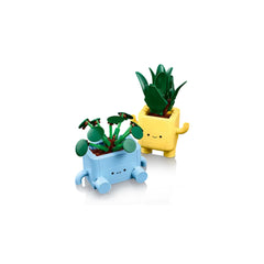 Two colorful LEGO potted plants with smiling faces, featuring a light blue baby dracaena and a yellow baby pilea, both with vibrant green leaves.