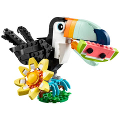 Colorful LEGO toucan with a black body and vibrant beak, holding a watermelon slice, perched on a green base with a yellow flower.