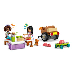 Two LEGO Friends characters, Liann and Aliya, seated at a table with fruits and drinks, preparing for a road trip. A small puppy figure sits beside them, while a colorful travel car with a trailer is parked nearby, completing this playful scene.