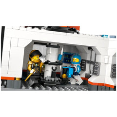 Two LEGO minifigures inside a space base: one in a black and gold suit with a staff, the other in a blue suit holding a cup, surrounded by control panels and equipment.