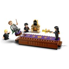 Four LEGO minifigures stand around a dueling platform. A blonde figure in green robes is airborne, while another figure aims a wand at him. To the left, a dark-robed character points, and in the center, a boy with glasses watches. A small ornate structure with a flame is featured in the background, enhancing the magical atmosphere of this Harry Potter-themed playset.