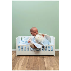 Doll in a white wooden cradle with blue bedding, holding a soft toy, against a mint green wall.