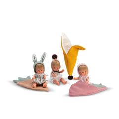 Three ASI Dolls seated, each with blonde hair and colorful hats, accompanied by whimsical, textured blankets and a yellow toy.