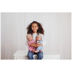 Child holding two realistic dolls, one in pink pajamas, one in blue pajamas, both wearing headbands, seated on a white table.