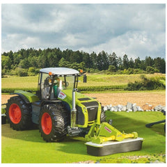 Claas toy tractor with front disc mower on green field, featuring realistic details and vibrant colors, against a forest backdrop.