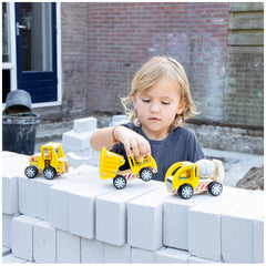 Child with blonde hair plays with yellow and red toy construction vehicles on a concrete ground against a brick wall.