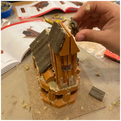 Teifoc Church model being constructed by a person's hand, showcasing light and dark brown wooden planks on a flat surface.
