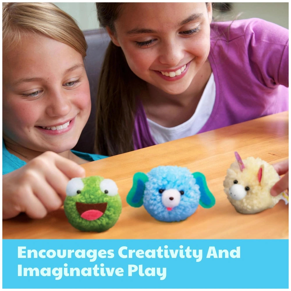 4M Make Your Own Pom Pom kit displayed on a brown wooden tabletop with two smiling girls in purple shirts in the background.