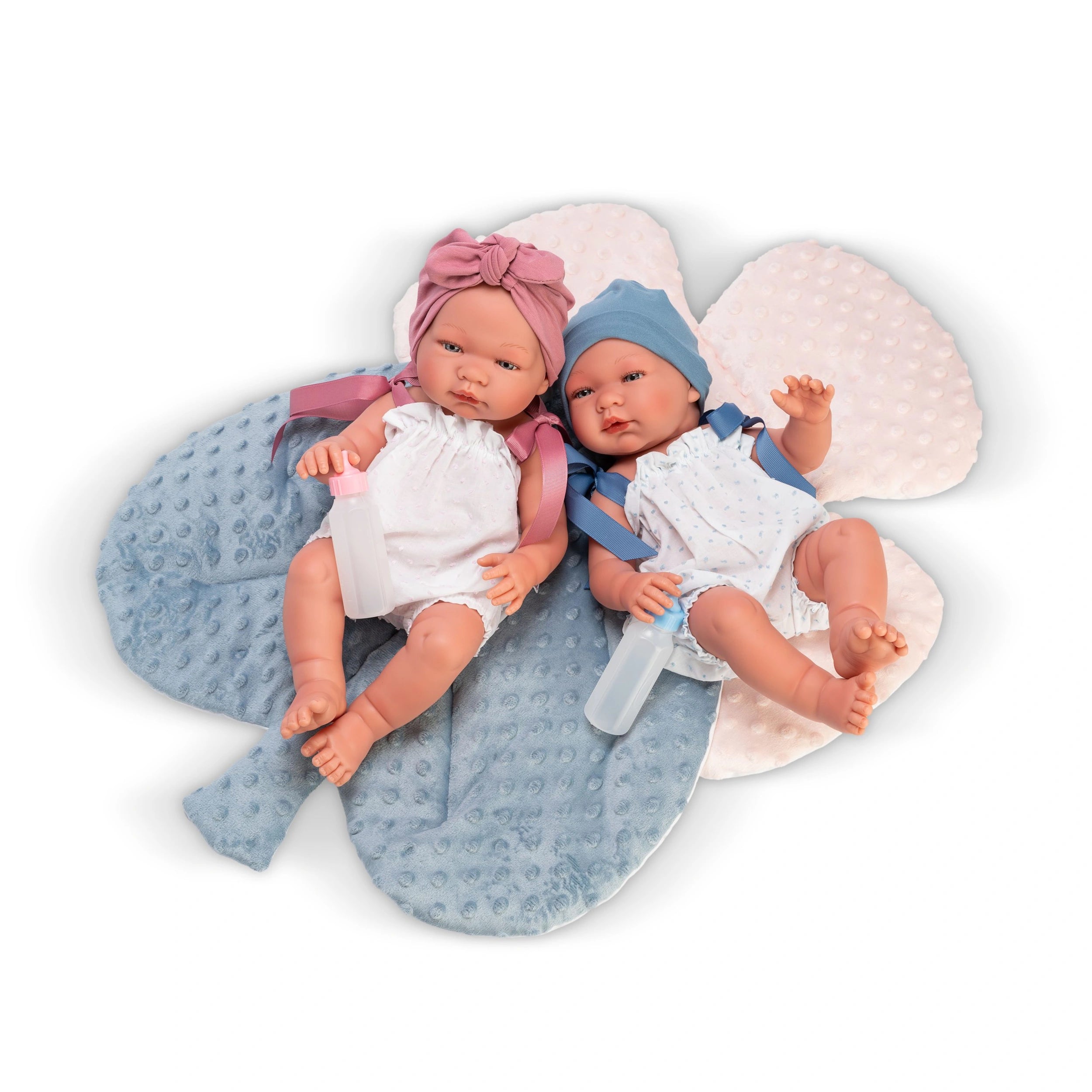 Two ASI Dolls, one with a pink headband and the other with a blue hat, lie on a textured leaf pillow with bottles.
