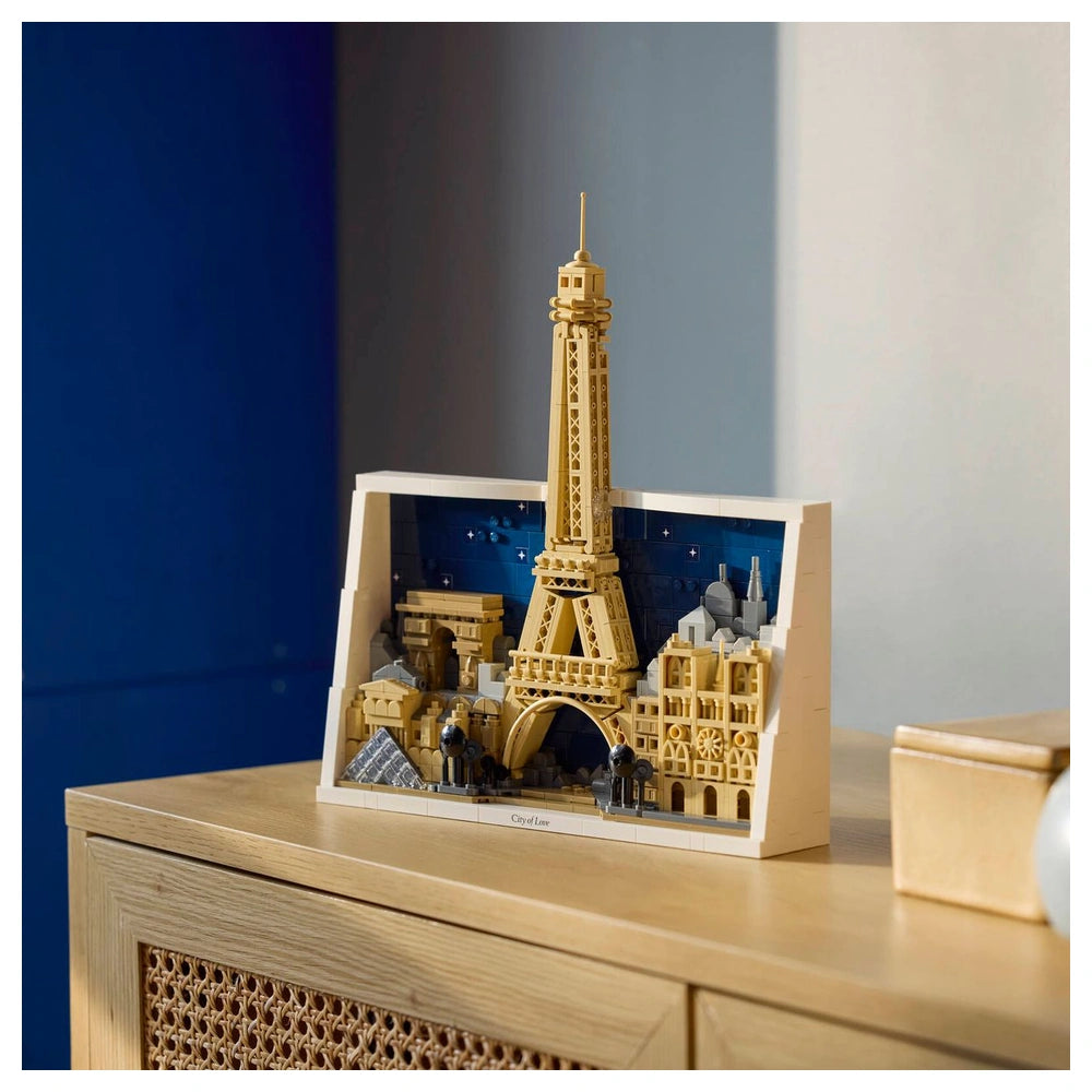 LEGO Eiffel Tower model in yellow on a wooden surface, set against a light wall and a simplified cityscape backdrop.