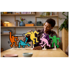 A smiling young man interacts with colorful LEGO figures inspired by Keith Haring, showcasing vibrant dancing poses, in a cozy room with wooden shelves and plants.