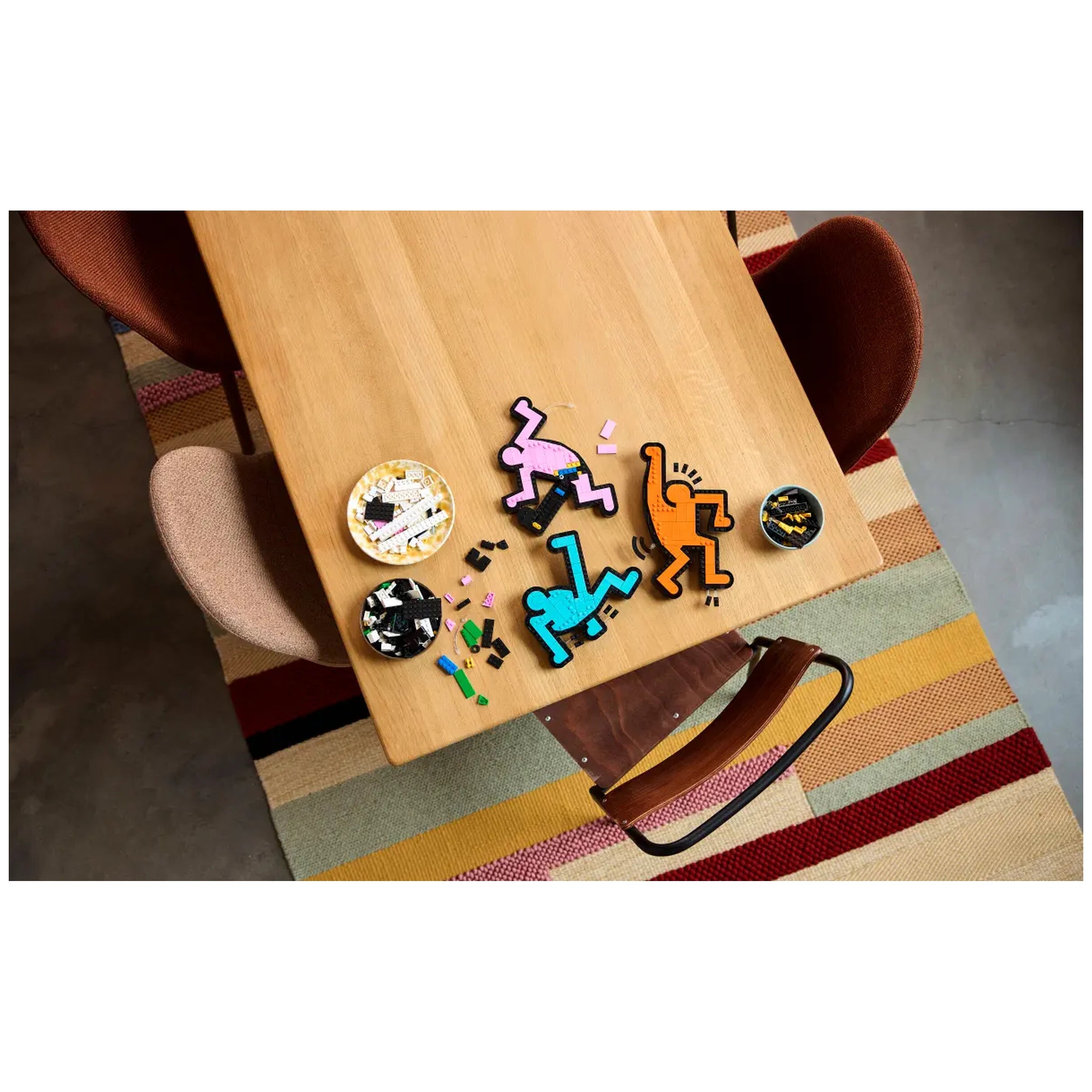 Colorful LEGO pieces arranged on a wooden table, featuring dancing figures inspired by Keith Haring's artwork, with crafting elements spread around.
