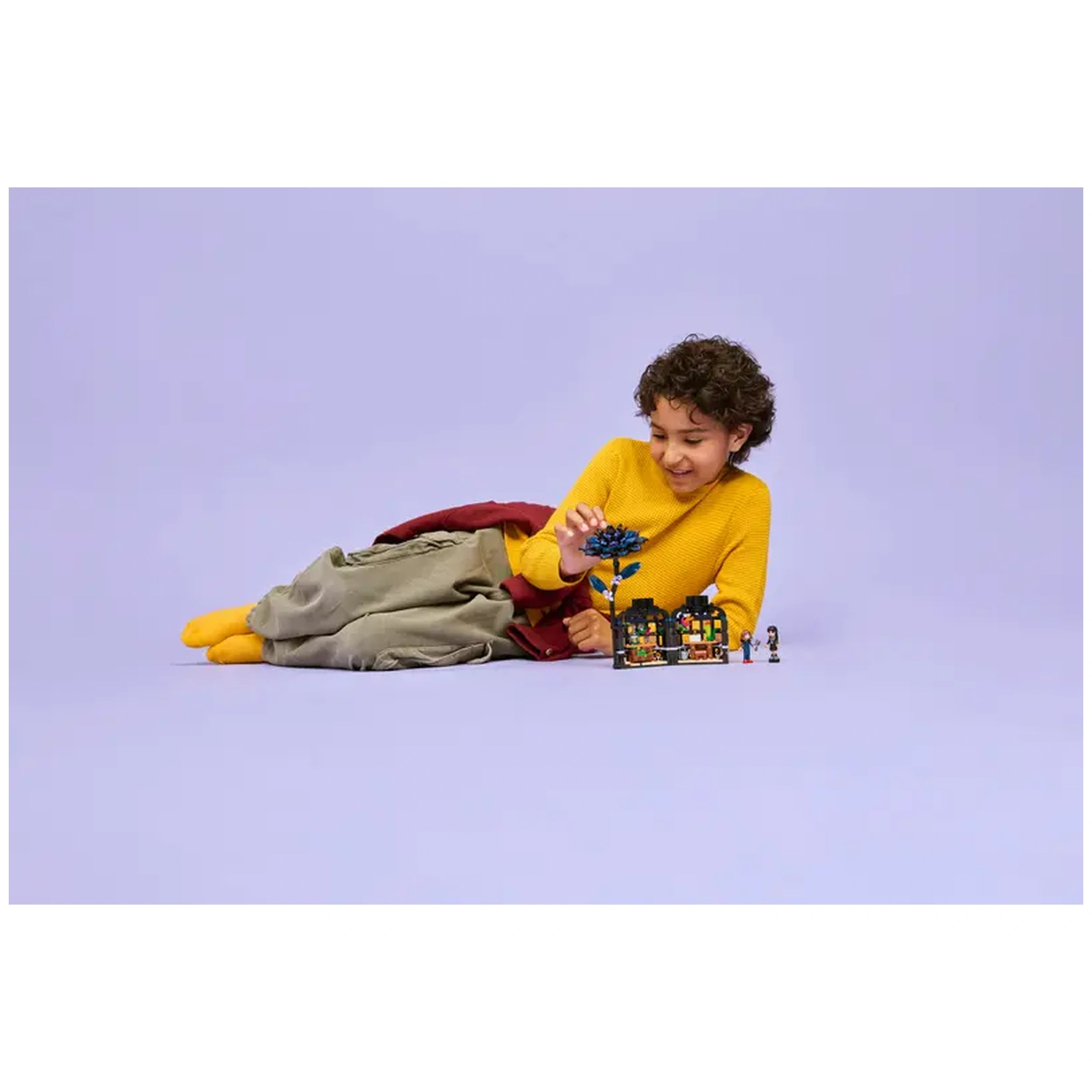 A child with curly hair, wearing a yellow sweater, happily interacts with the LEGO Black Dahlia Flower set, lying on a light purple background.