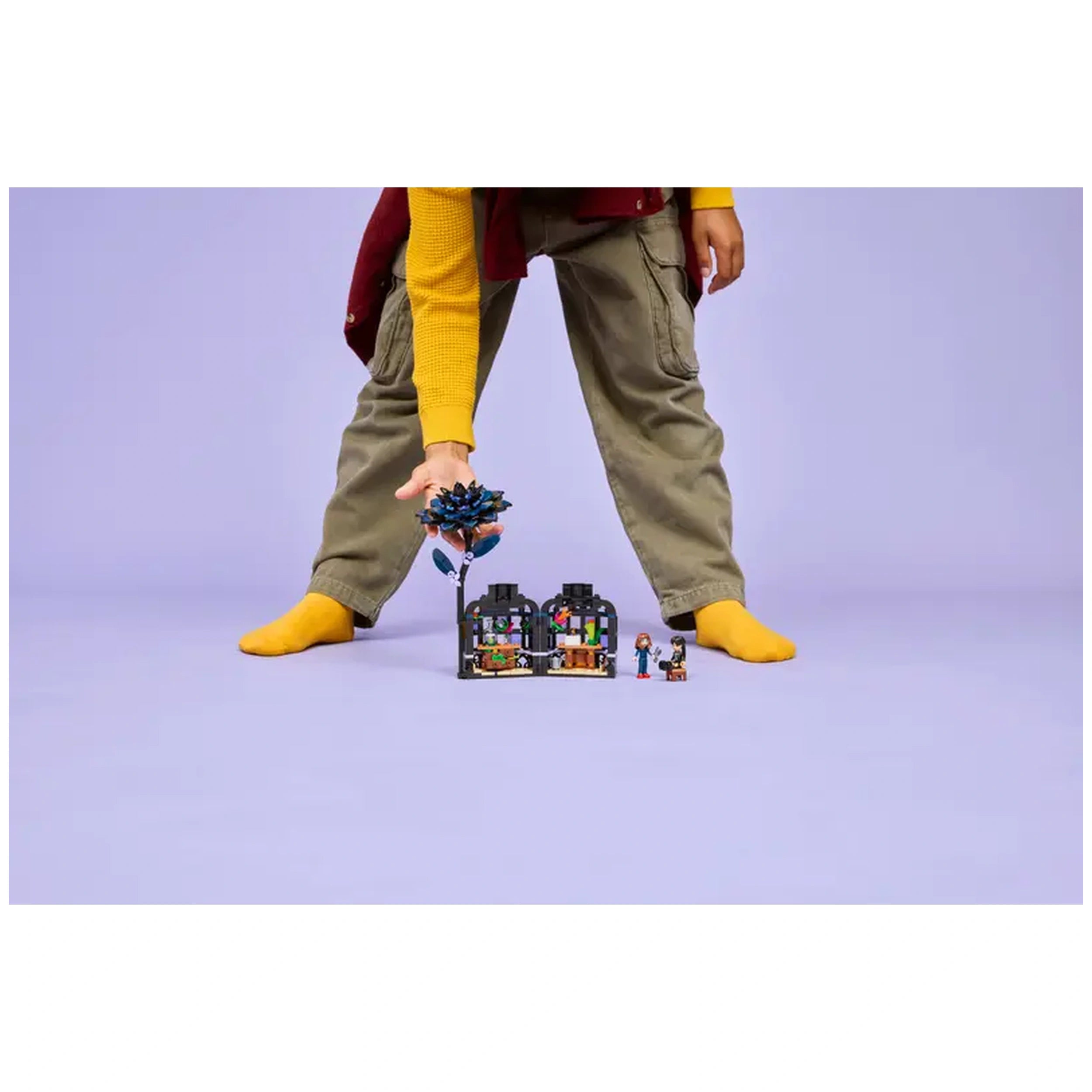 A child in a yellow sweater and gray pants holds a blue-black dahlia flower from the LEGO Black Dahlia Flower set, standing over a mini classroom scene. The background is a soft purple hue.