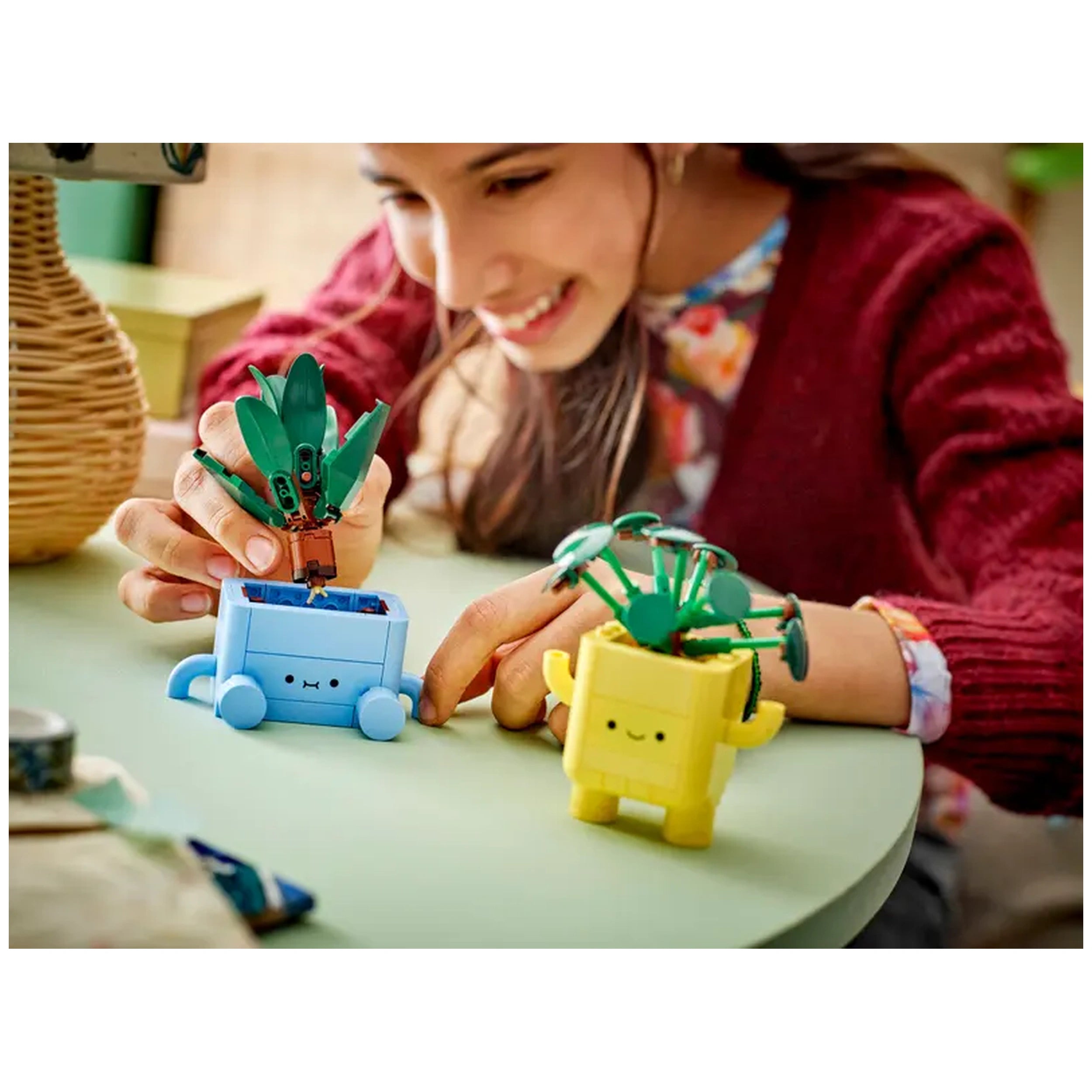 A child joyfully assembles colorful LEGO botanical plants, focusing on a blue pot with a baby dracaena while a cheerful yellow pot with a pilea sits beside her on a light green table.