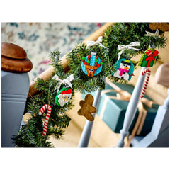 Four colorful LEGO® Christmas ornaments hang from a green garland on a staircase. The ornaments include a jolly Santa, a cheerful snowman, a playful reindeer, and a beautifully wrapped gift, all tied with festive bows. A striped candy cane and a gingerbread man add to the holiday decor.