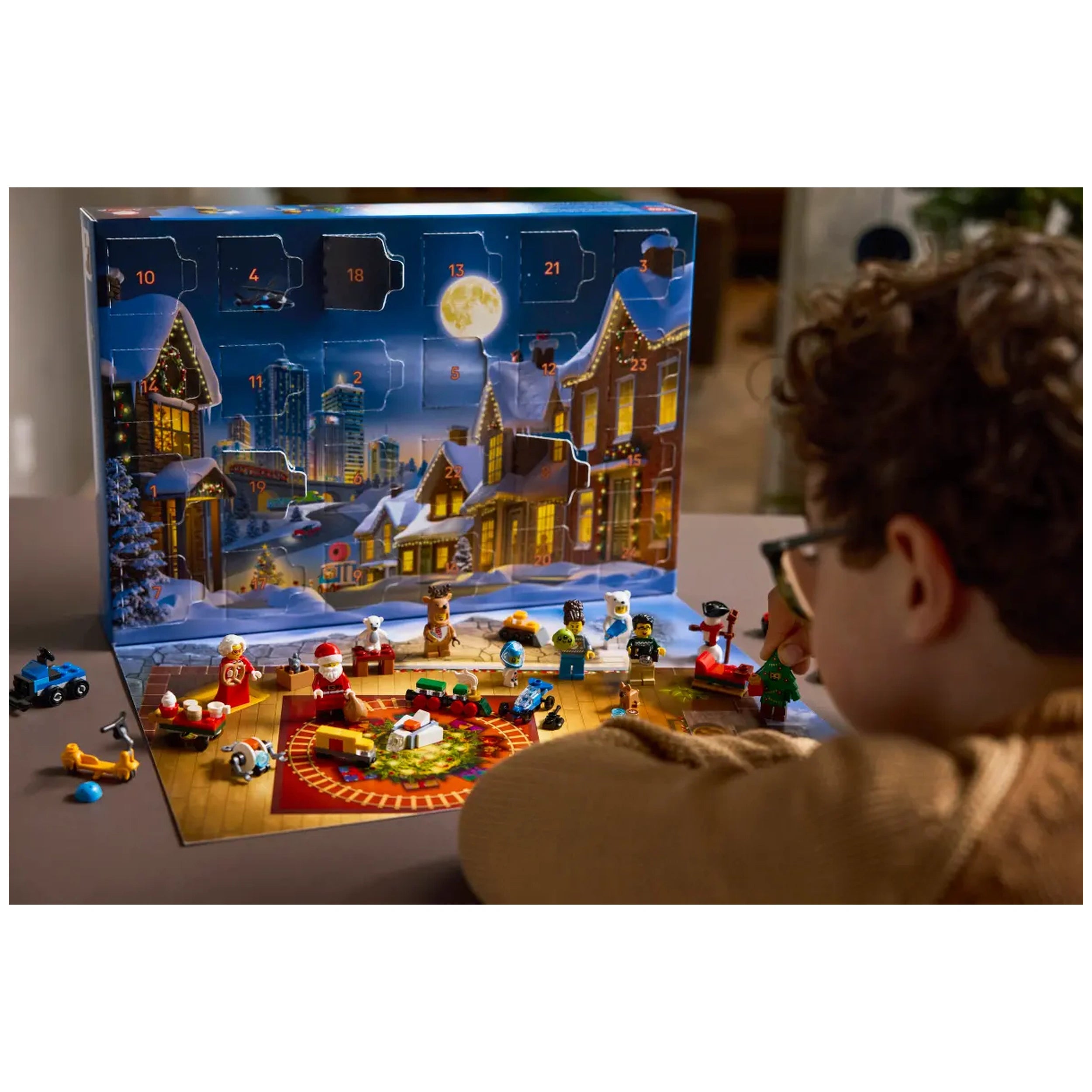 A child in a brown sweater engages with the LEGO City Advent Calendar 2025, surrounded by festive mini-builds and characters. The vibrant scene features a snowy background with charming decorated houses and a large, bright moon. Colorful accessories and toys are displayed on a playmat with a round festive carpet, enhancing holiday spirit and imaginative play.