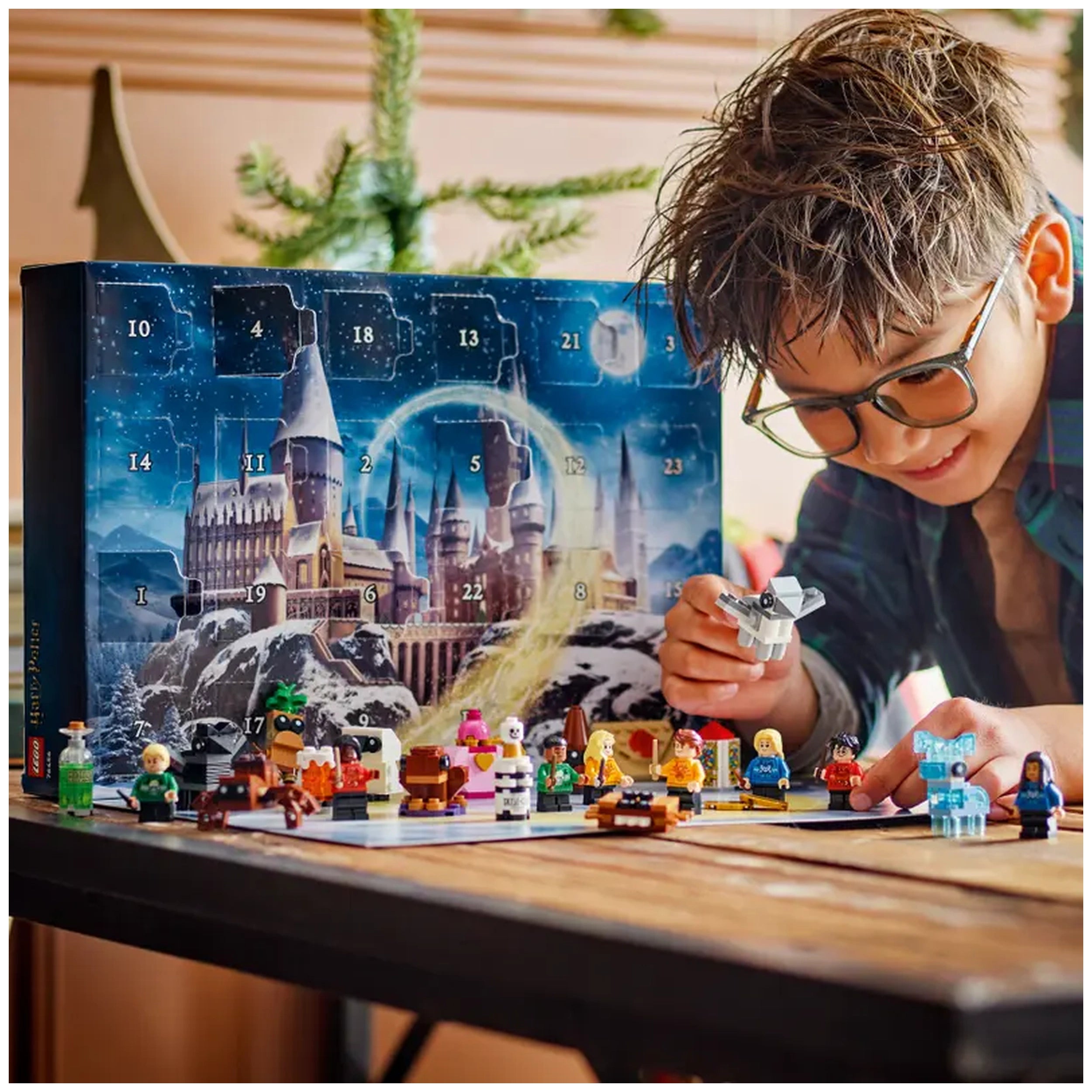 A joyful child in glasses interacts with the LEGO Harry Potter Advent Calendar on a wooden table. The colorful calendar features the Hogwarts castle and numbered doors. Surrounding the child are various buildable figures and festive minifigures, including Harry Potter and friends, all in holiday attire. Soft holiday decorations are visible in the background, creating a festive atmosphere.