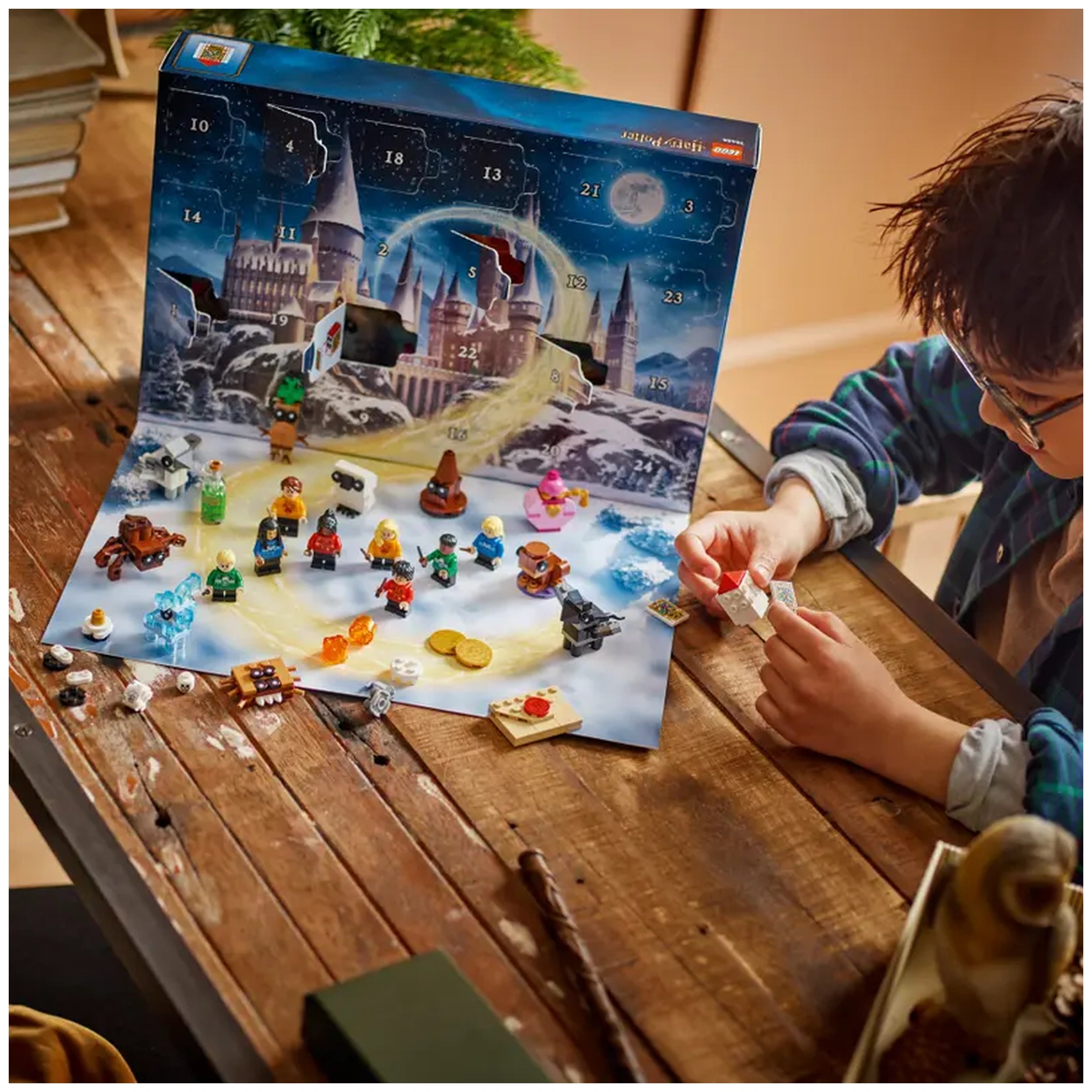 LEGO Harry Potter Advent Calendar 2025 displayed on a wooden table, showcasing colorful minifigures and buildable pieces against a snowy, Hogwarts-themed backdrop. A child interacts with the calendar, excitedly unveiling hidden surprises from the numbered doors.