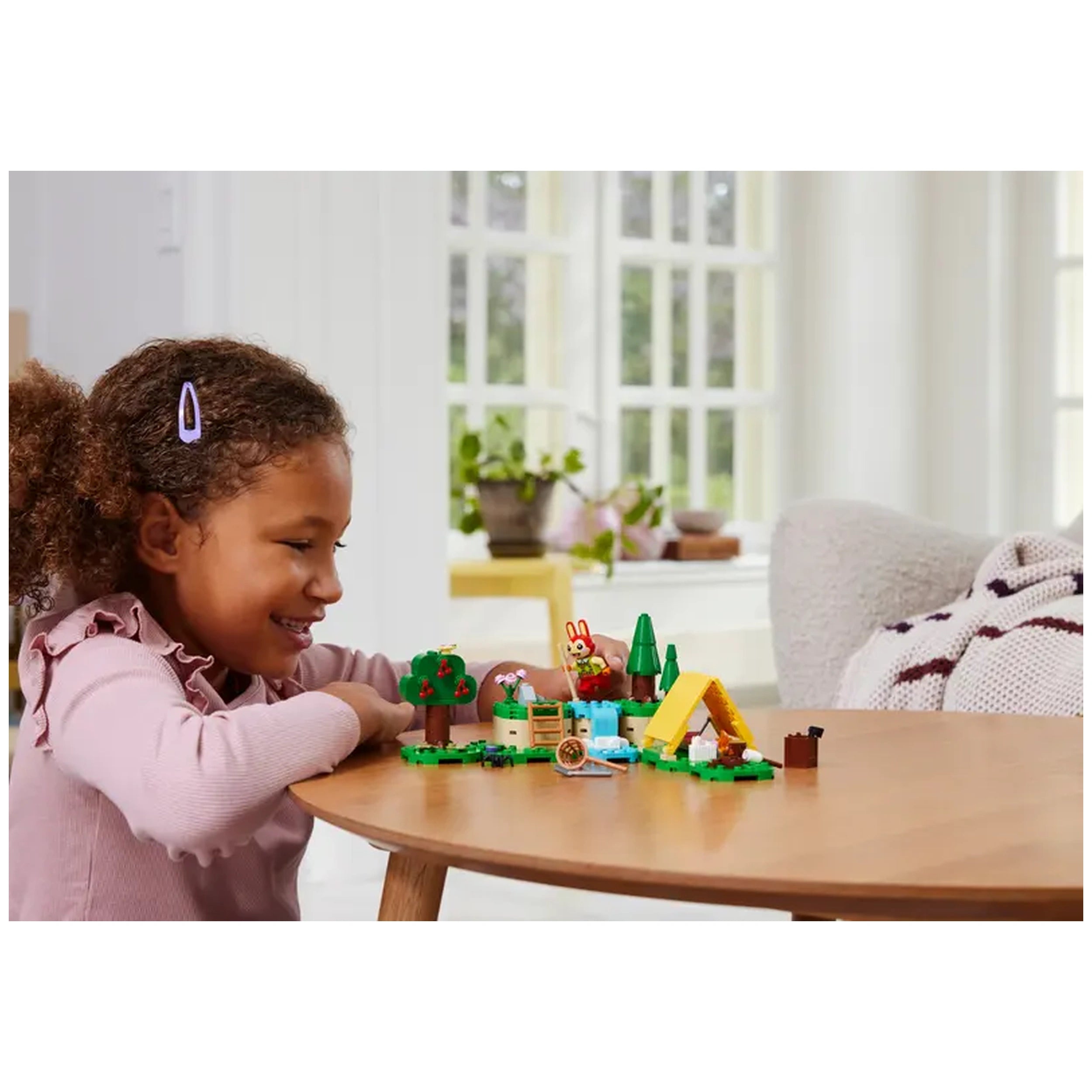 A young girl with curly hair smiles while playing at a wooden table with the LEGO Animal Crossing Bunnie's Outdoor Activities set, featuring a colorful campsite, trees, and a tent.