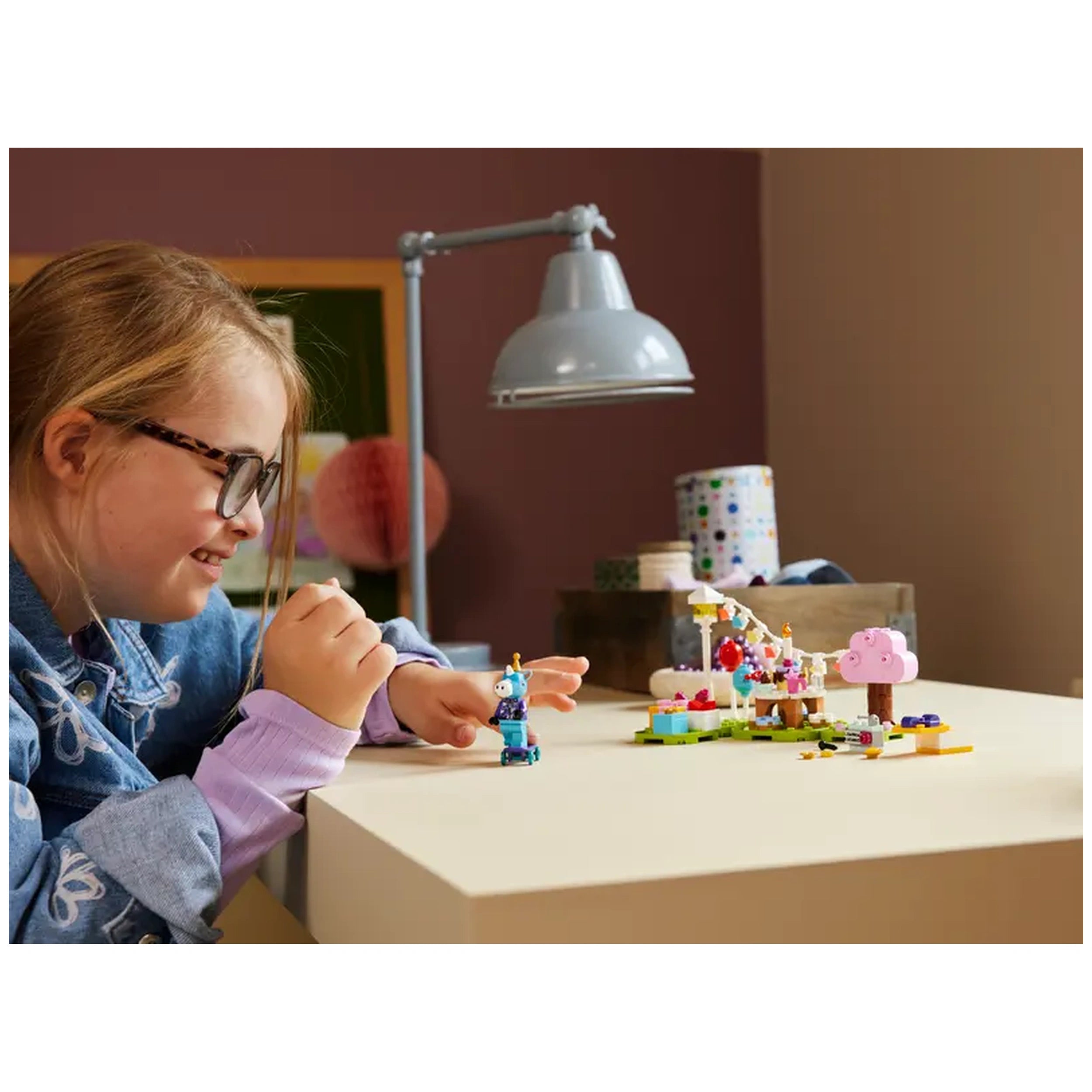 A child with glasses smiles while assembling the LEGO® Animal Crossing™ Julian's Birthday Party set on a light wooden table. Colorful decorations and a birthday cake with candles are visible, creating a festive scene. A desk lamp illuminates the cheerful setup in a cozy room.