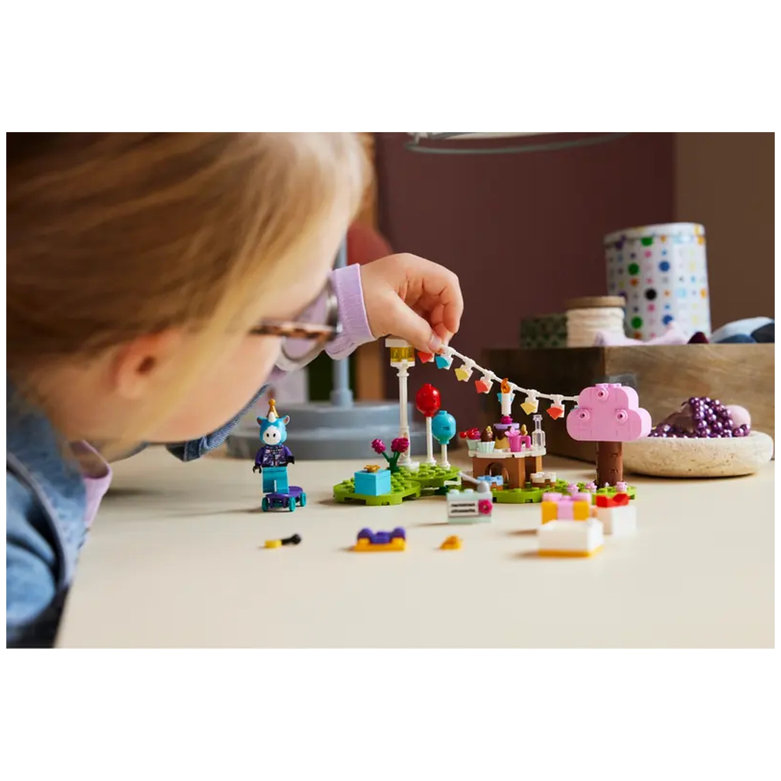 A child with glasses joyfully arranges colorful LEGO® pieces for Julian’s Birthday Party, featuring a pink tree, balloons, and a cake on a tabletop.