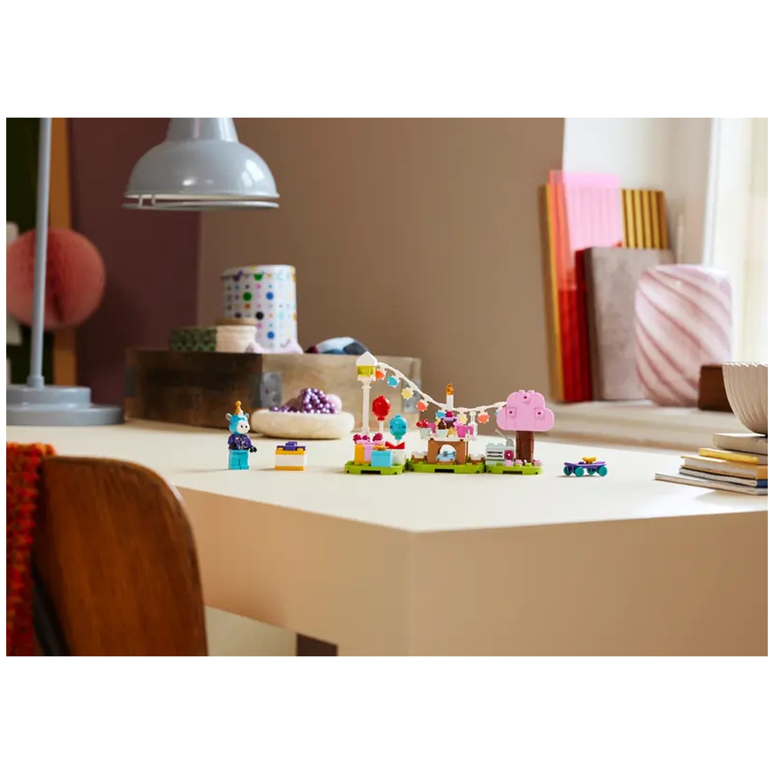 Colorful LEGO® Animal Crossing™ Julian’s Birthday Party set displayed on a light-colored table, featuring a birthday scene with a cake, gifts, and a character. A gray lamp and various craft supplies are in the background.