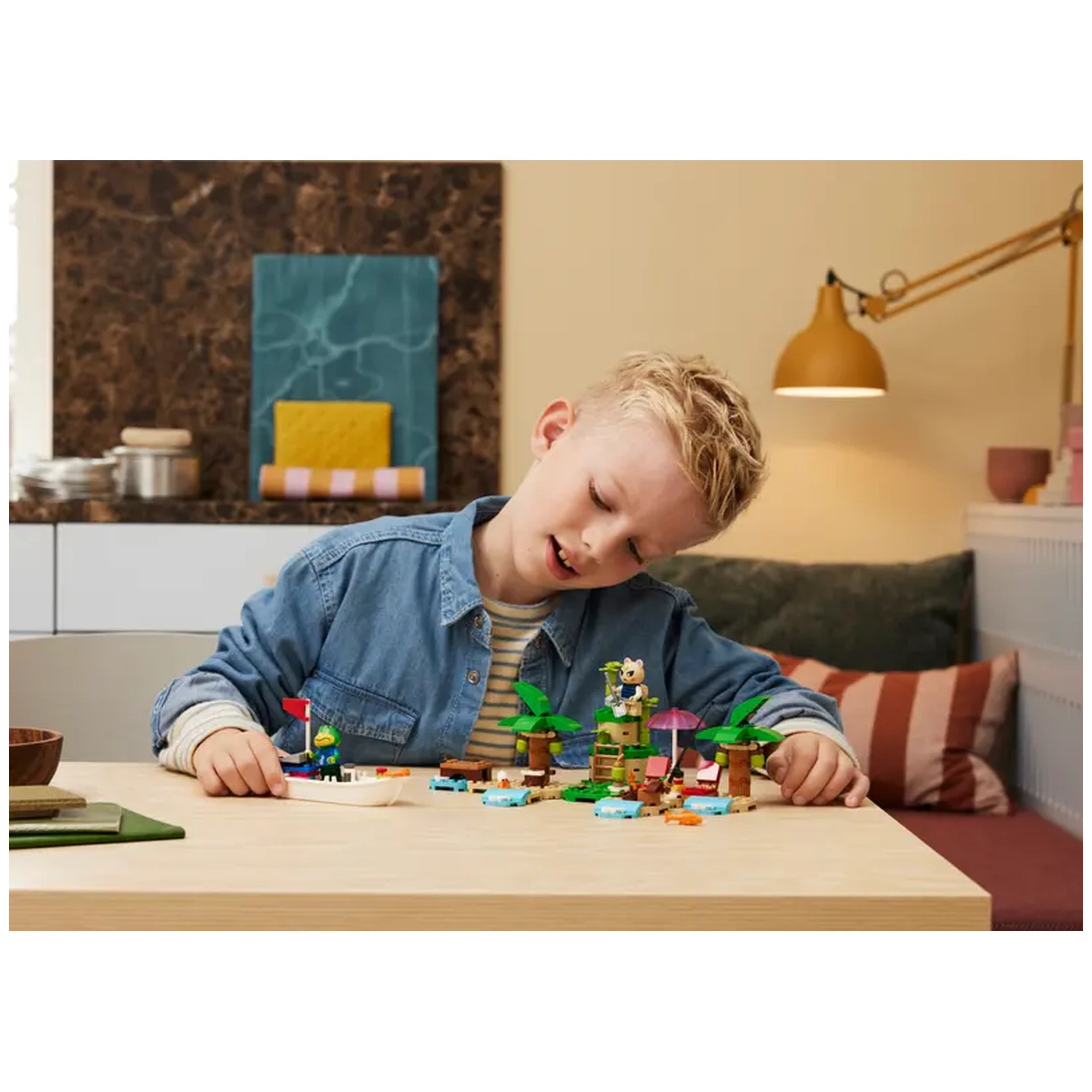 A young boy in a blue denim jacket engages with the LEGO Animal Crossing Kapp'n's Island Boat Tour set, surrounded by colorful building pieces, palm trees, and a small boat on a wooden table in a bright room with modern decor.