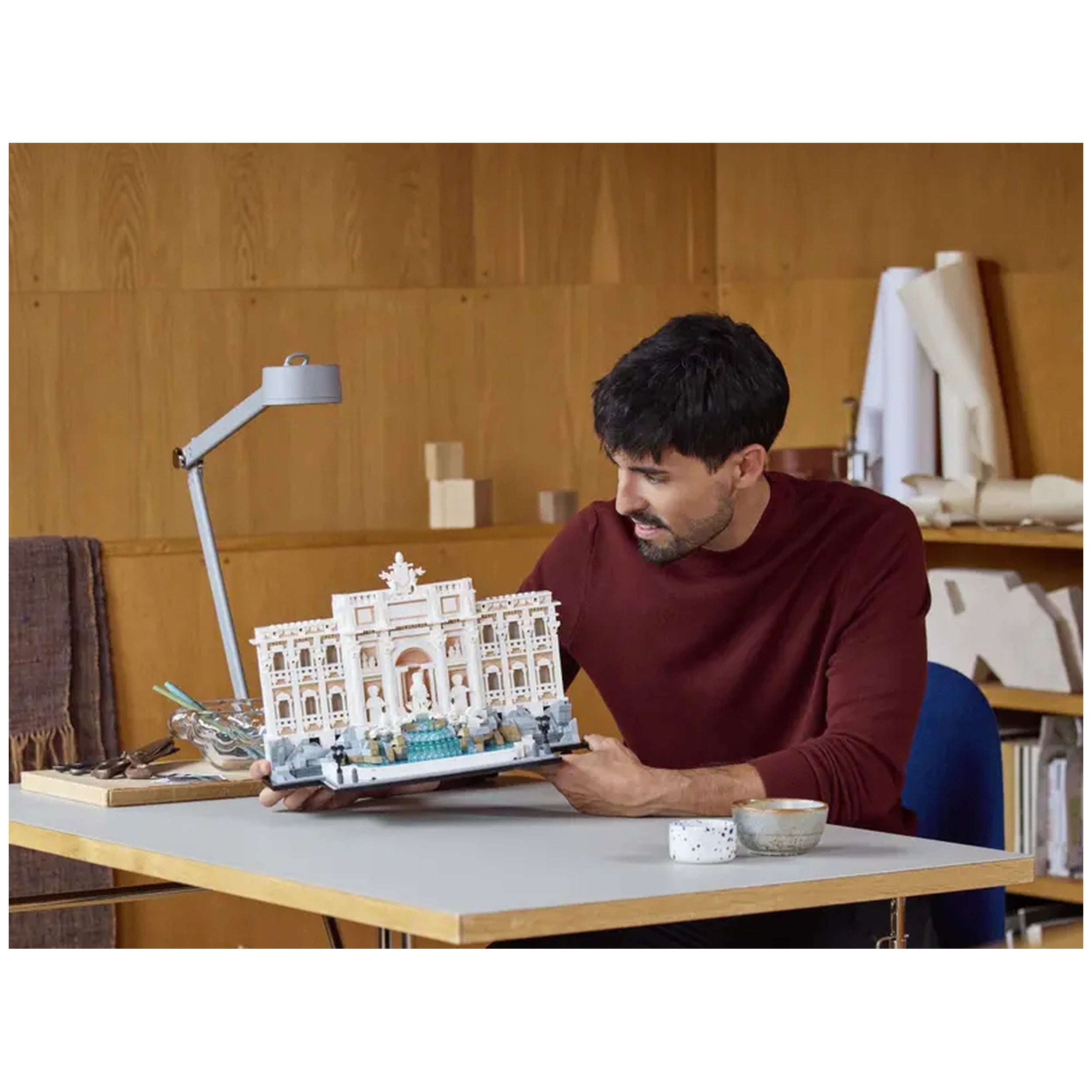 A man in a burgundy shirt joyfully admires a detailed LEGO replica of the Trevi Fountain, placed on a wooden table. Constructed with intricate elements, the model features the fountain's grandeur alongside a small bowl and cup. A modern lamp illuminates the workspace in a cozy, wooden-paneled room.