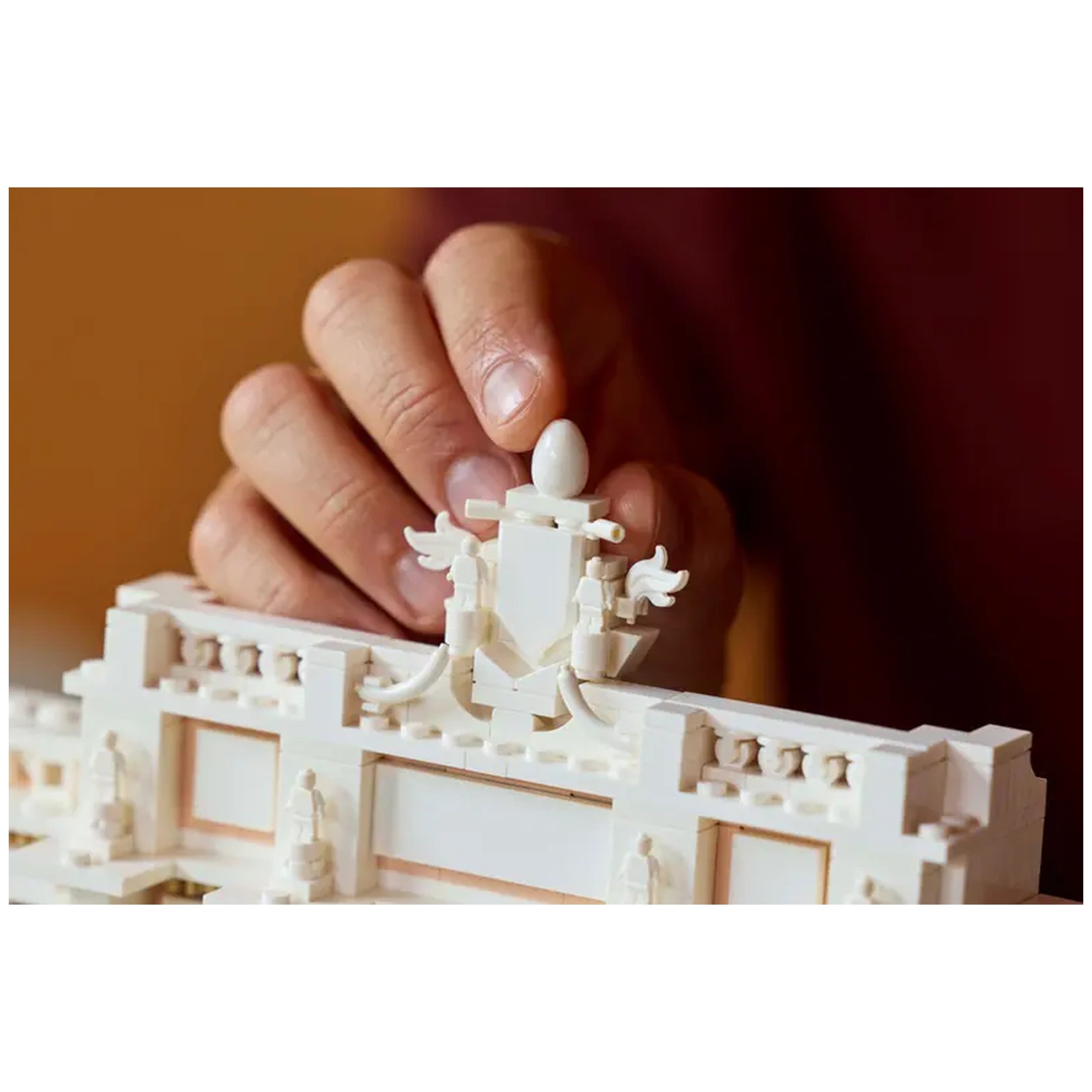 A person gently places a white LEGO® piece shaped like an egg atop an intricately designed section of the Trevi Fountain model, showcasing architectural details and statues.