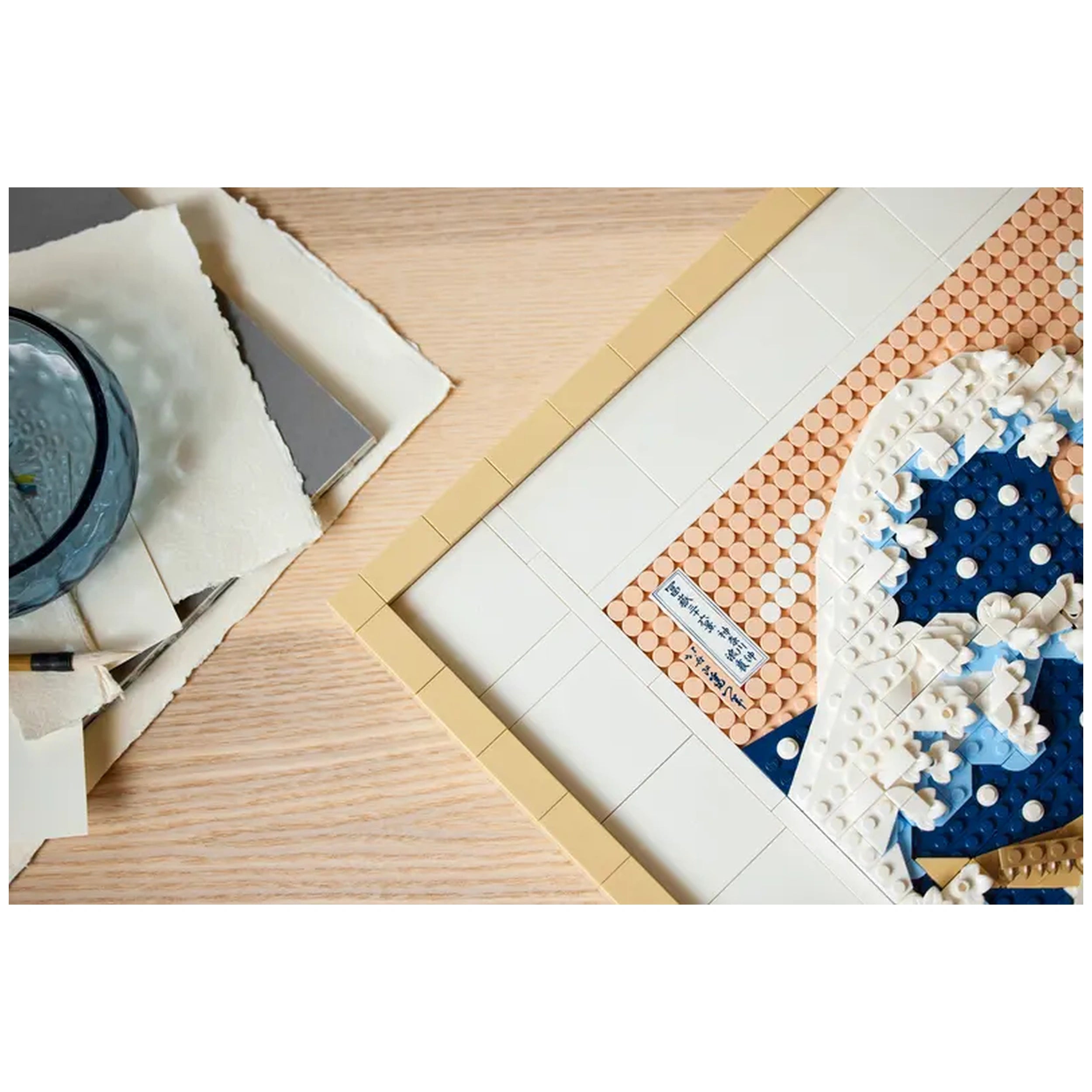 LEGO® Art set showcasing Hokusai's The Great Wave in progress, featuring vibrant blue and white pieces on a wooden surface. A glass of water and art materials are nearby.