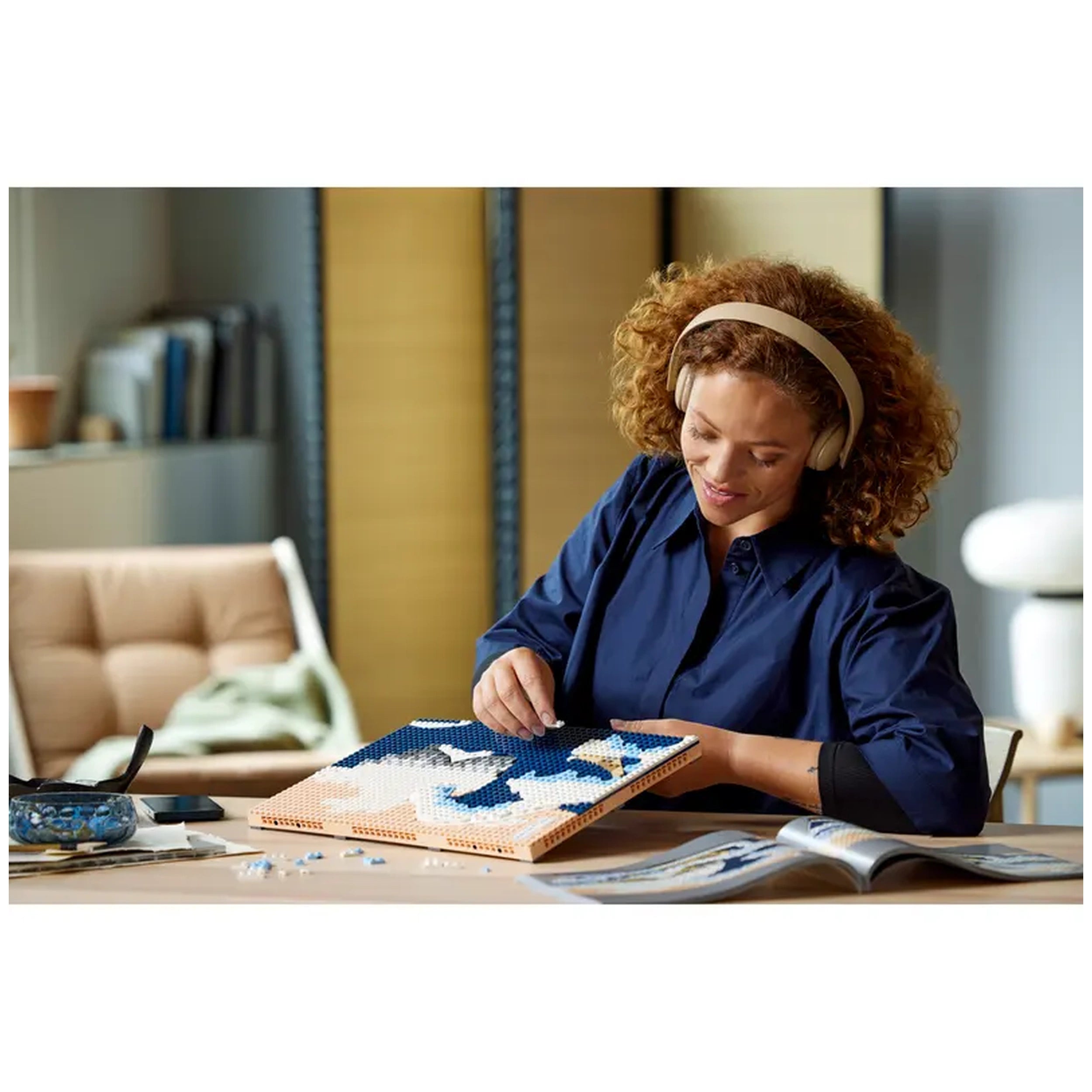 A woman with curly hair and headphones smiles while assembling a LEGO® Art set, featuring blue and white pieces, on a wooden table. Behind her, a cozy living space with a soft chair and decorative items is visible.
