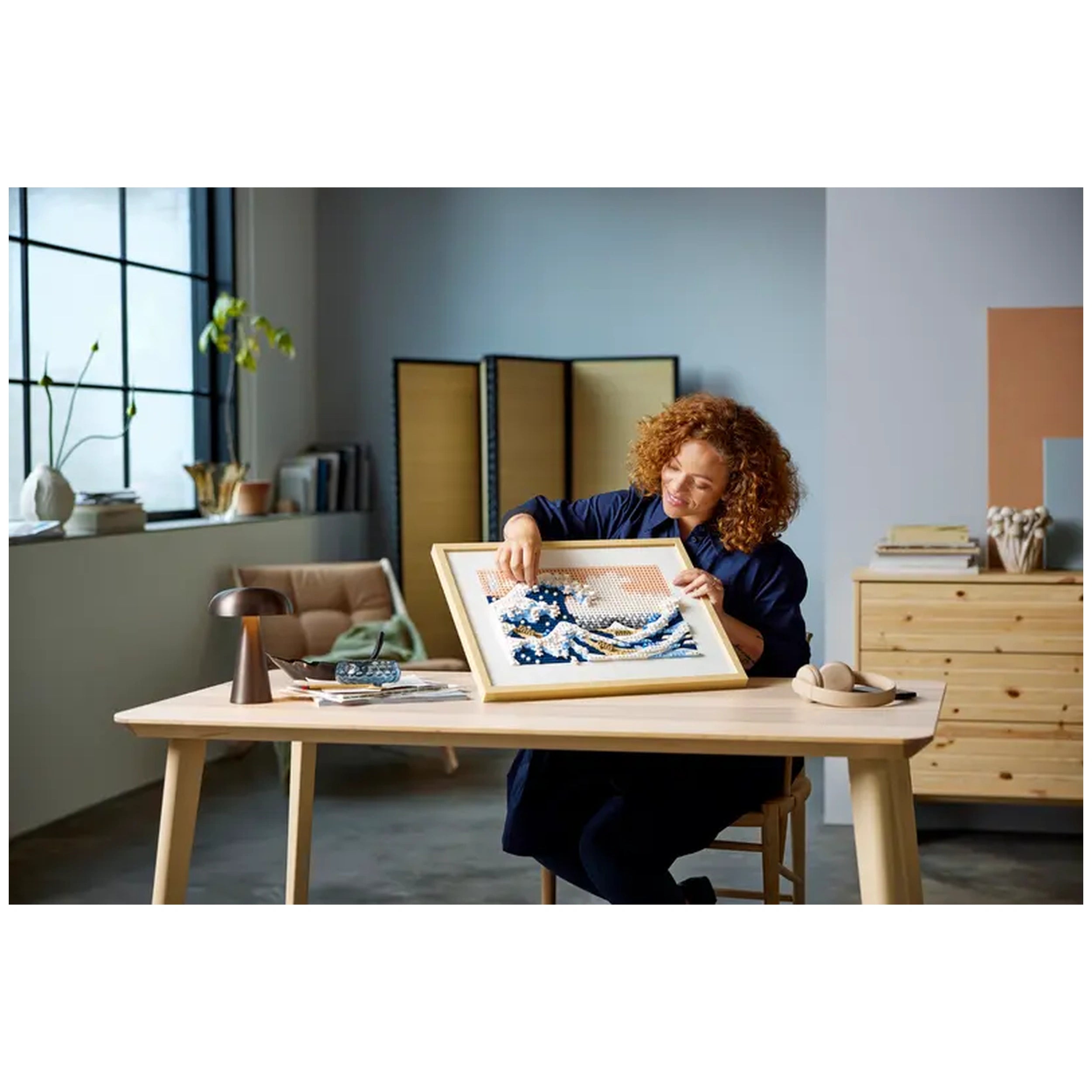 A woman with curly red hair is placing a framed LEGO® art piece of Hokusai’s The Great Wave on a wooden table. The stylish room features large windows, a plant, and minimalist decor.