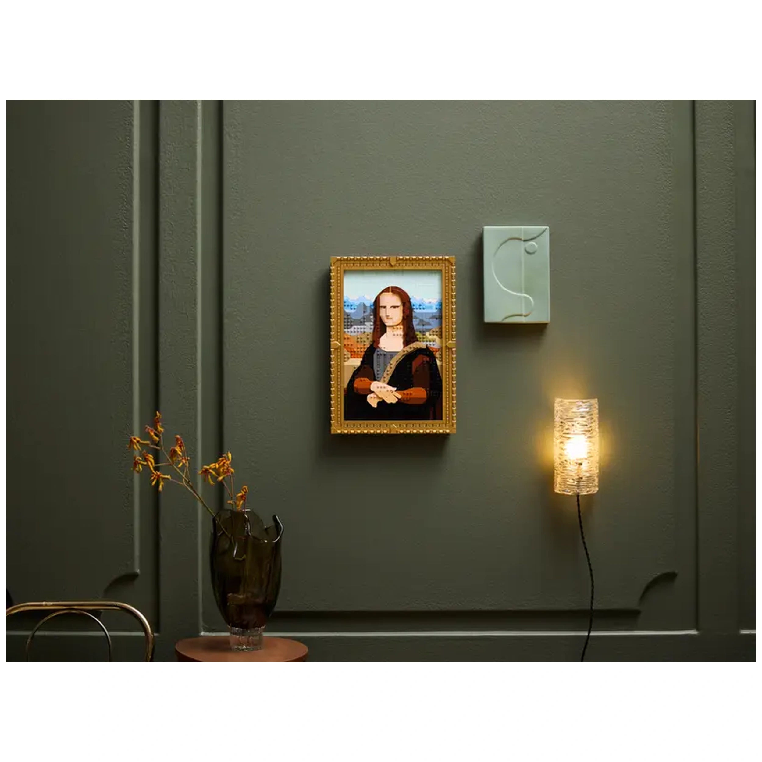 LEGO® Art Mona Lisa construction set displayed in a gold frame on a dark green wall, with a modern wall sconce and a vase of flowers nearby.