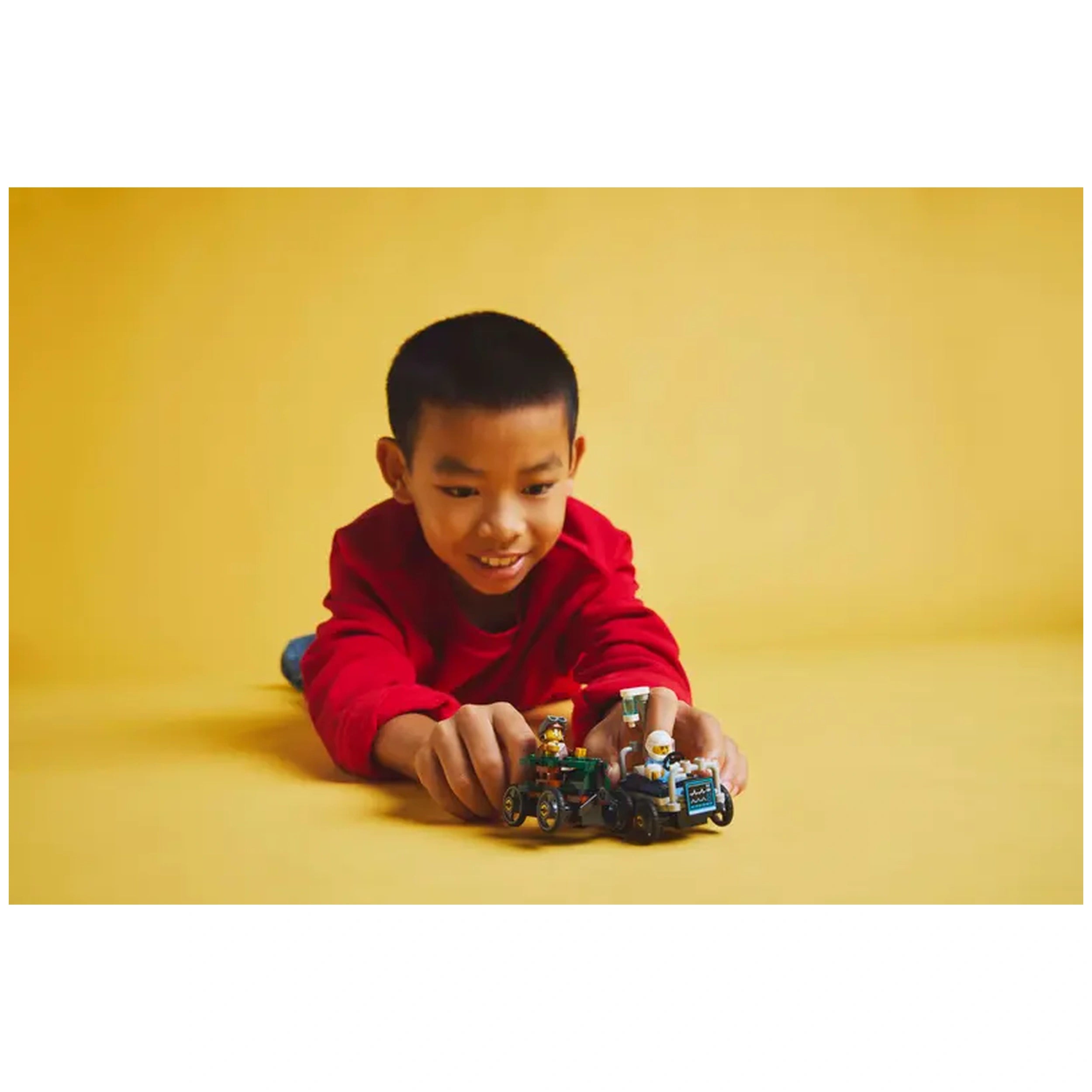 Young boy in a red sweatshirt excitedly plays on a yellow background with LEGO® race cars featuring a pilot and a hospital bed minifigure.