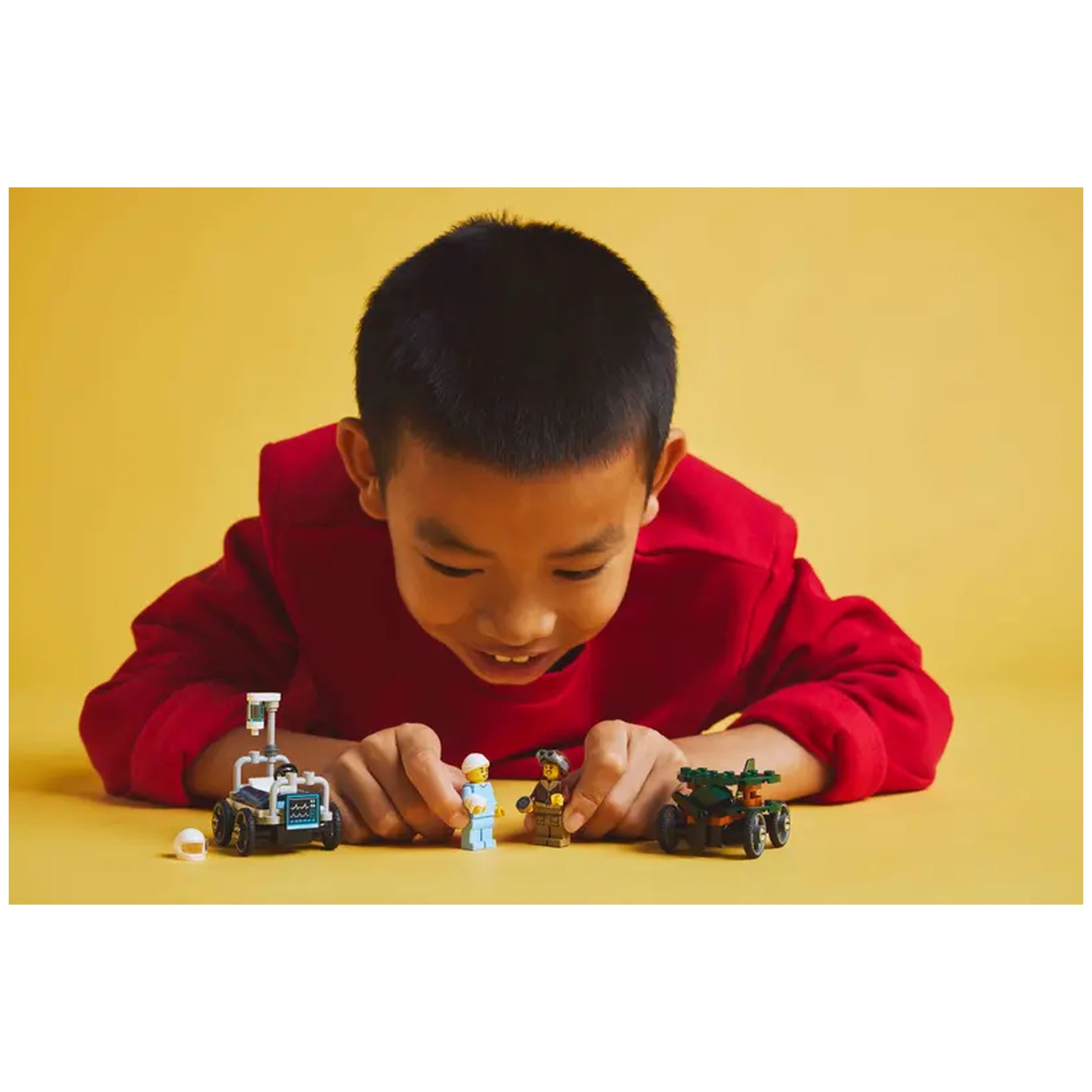 A young boy in a red sweatshirt smiles while playing with LEGO minifigures and soapbox race cars on a yellow background.