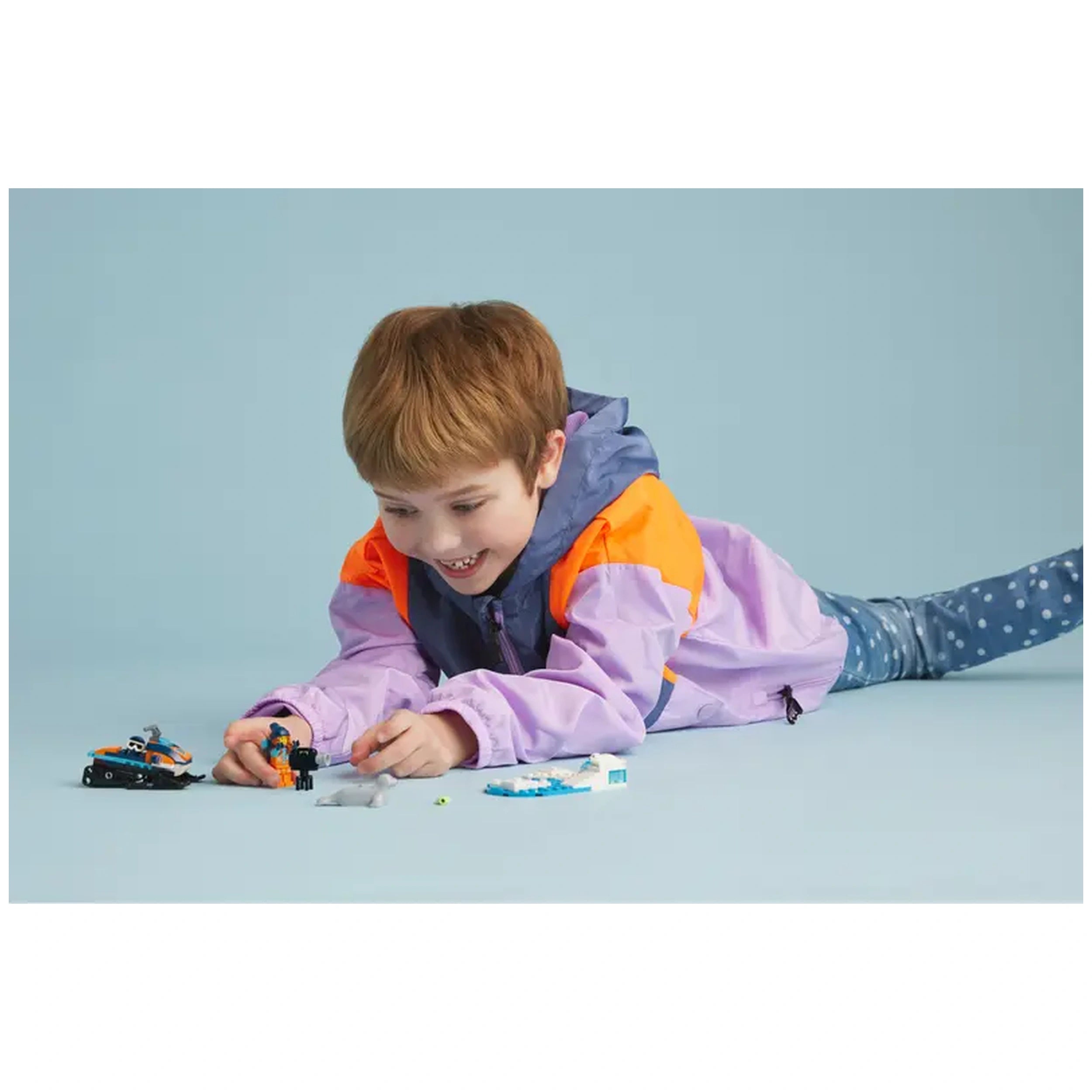 A young child with short brown hair plays on the floor, assembling the LEGO Arctic Explorer Snowmobile set. They are smiling, wearing a colorful purple and orange jacket, and surrounded by a small seal figure and building pieces. The background is a soft blue, creating a cheerful atmosphere for play.