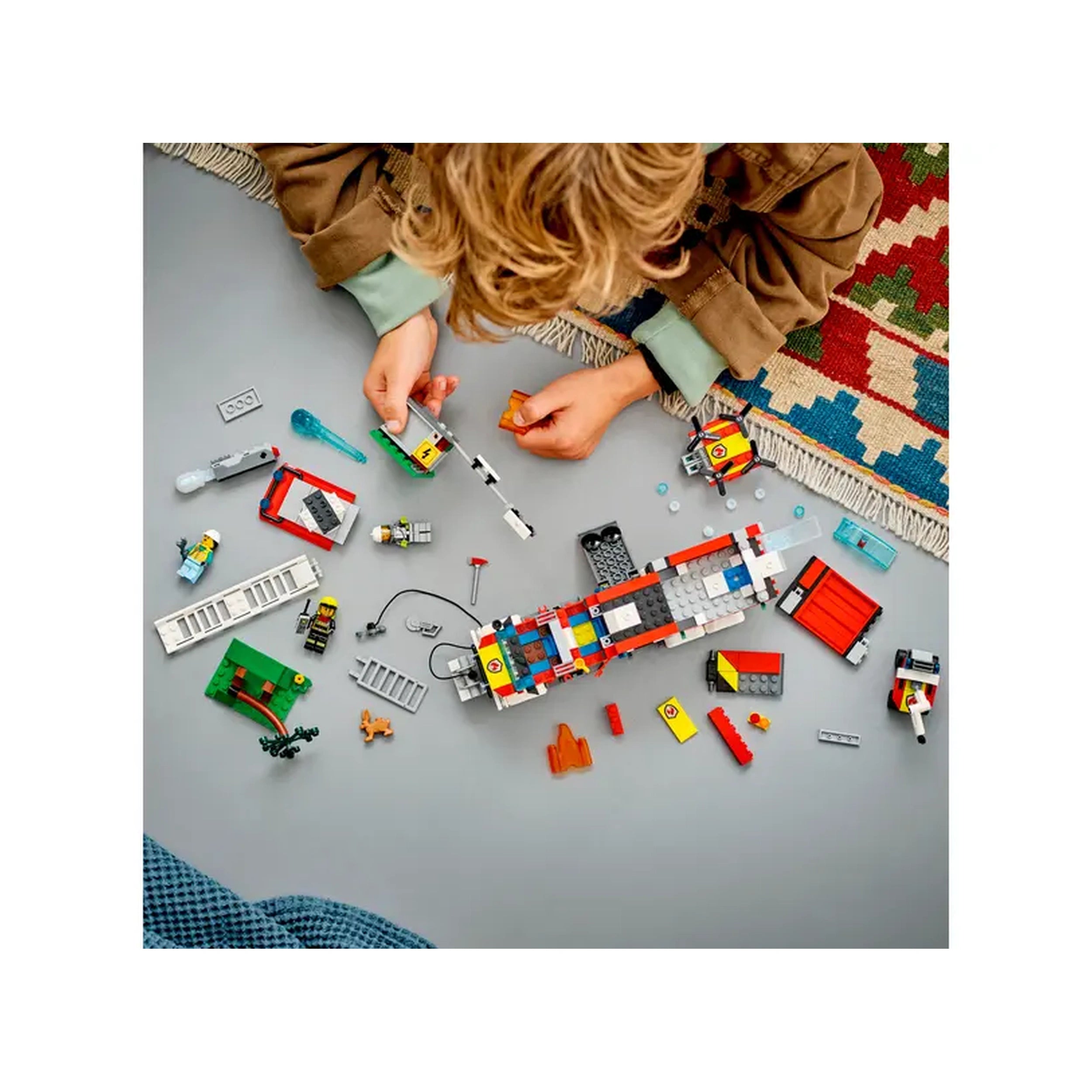 A child builds the LEGO® City Fire Command Truck set with various colored bricks, minifigures, and firefighting accessories scattered around on a gray surface.