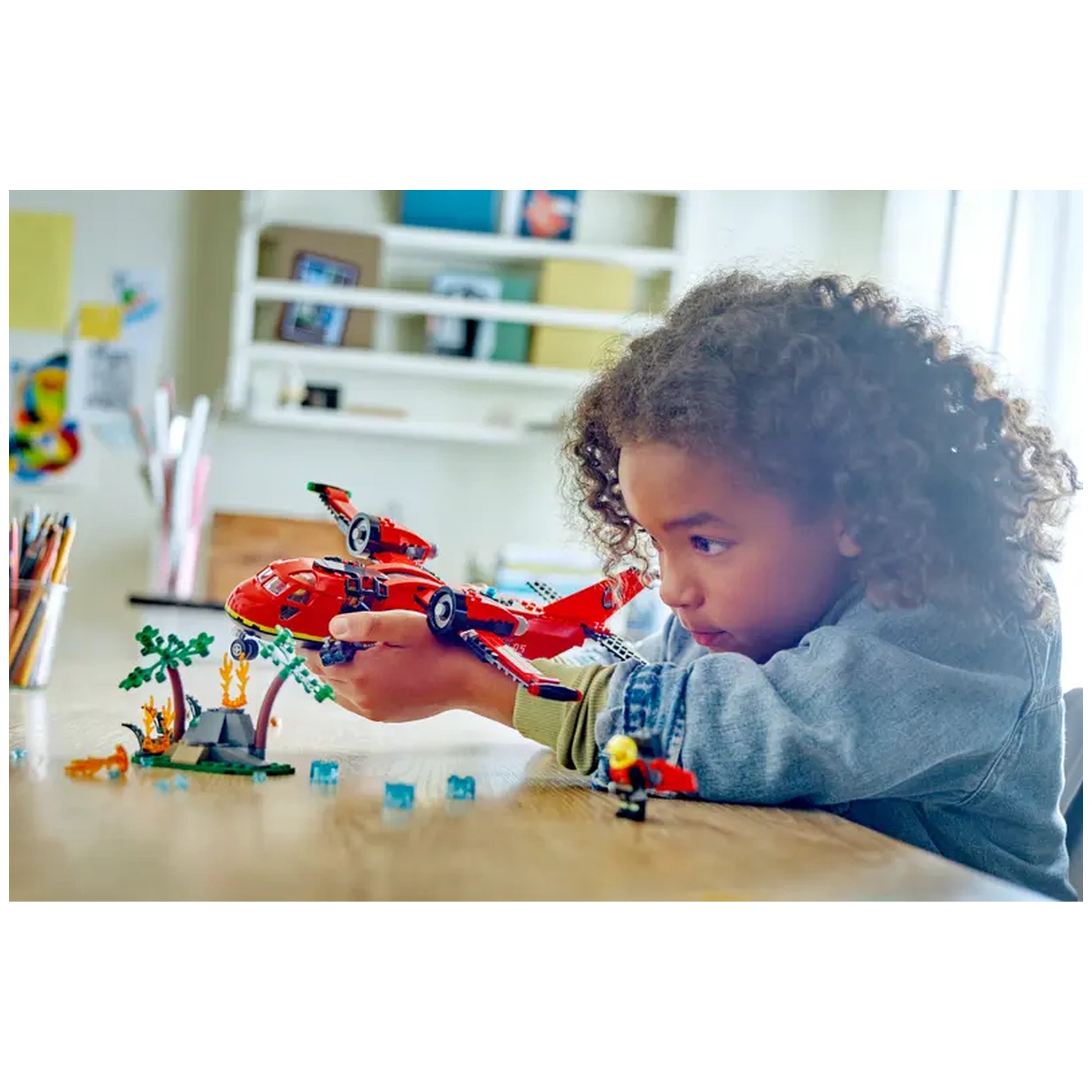 A child with curly hair plays with a red LEGO® City Fire Rescue Plane, concentrating on a scene with trees and minifigures on a wooden table.