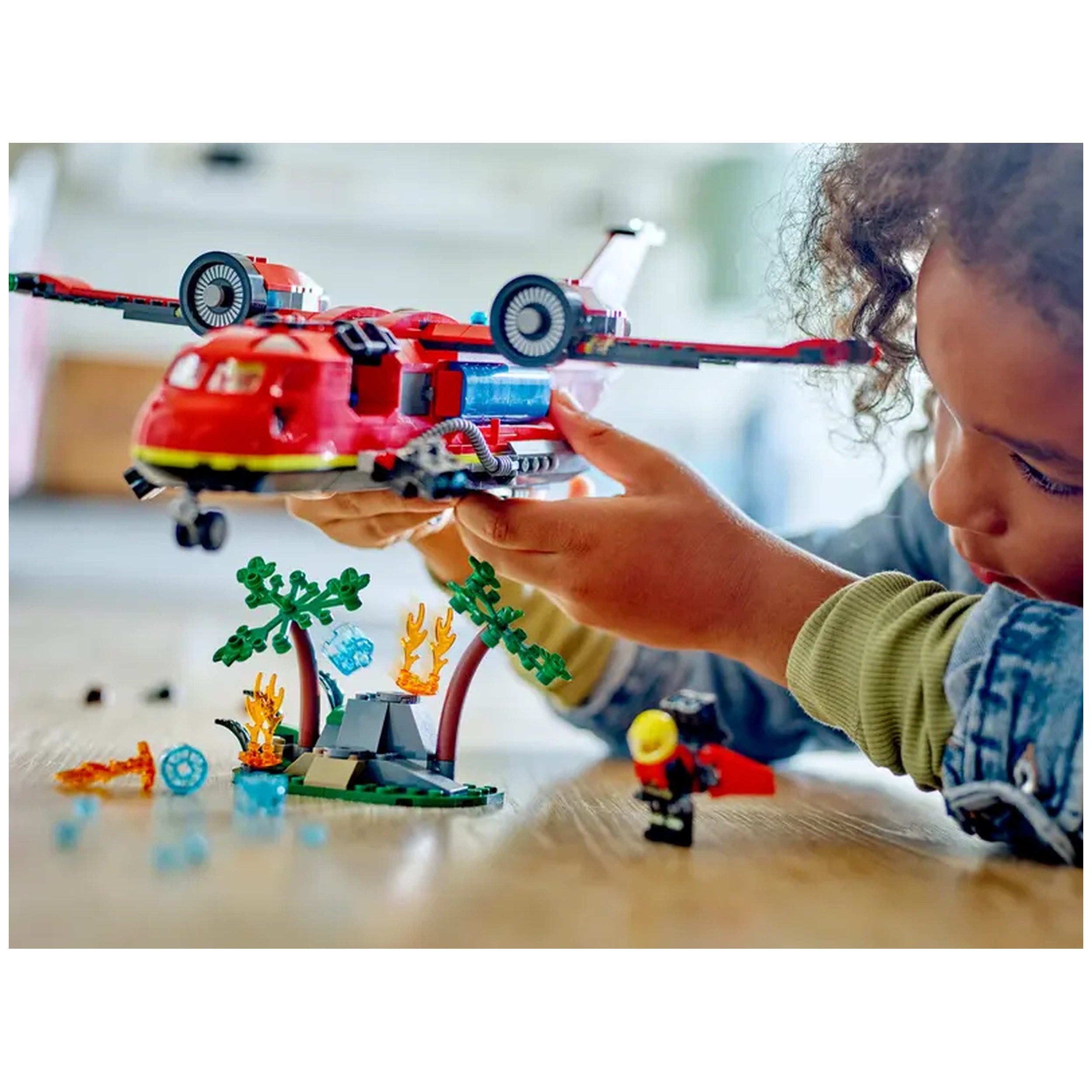 A child holds a red LEGO® City Fire Rescue Plane above a colorful firefighting scene featuring trees and fire elements.