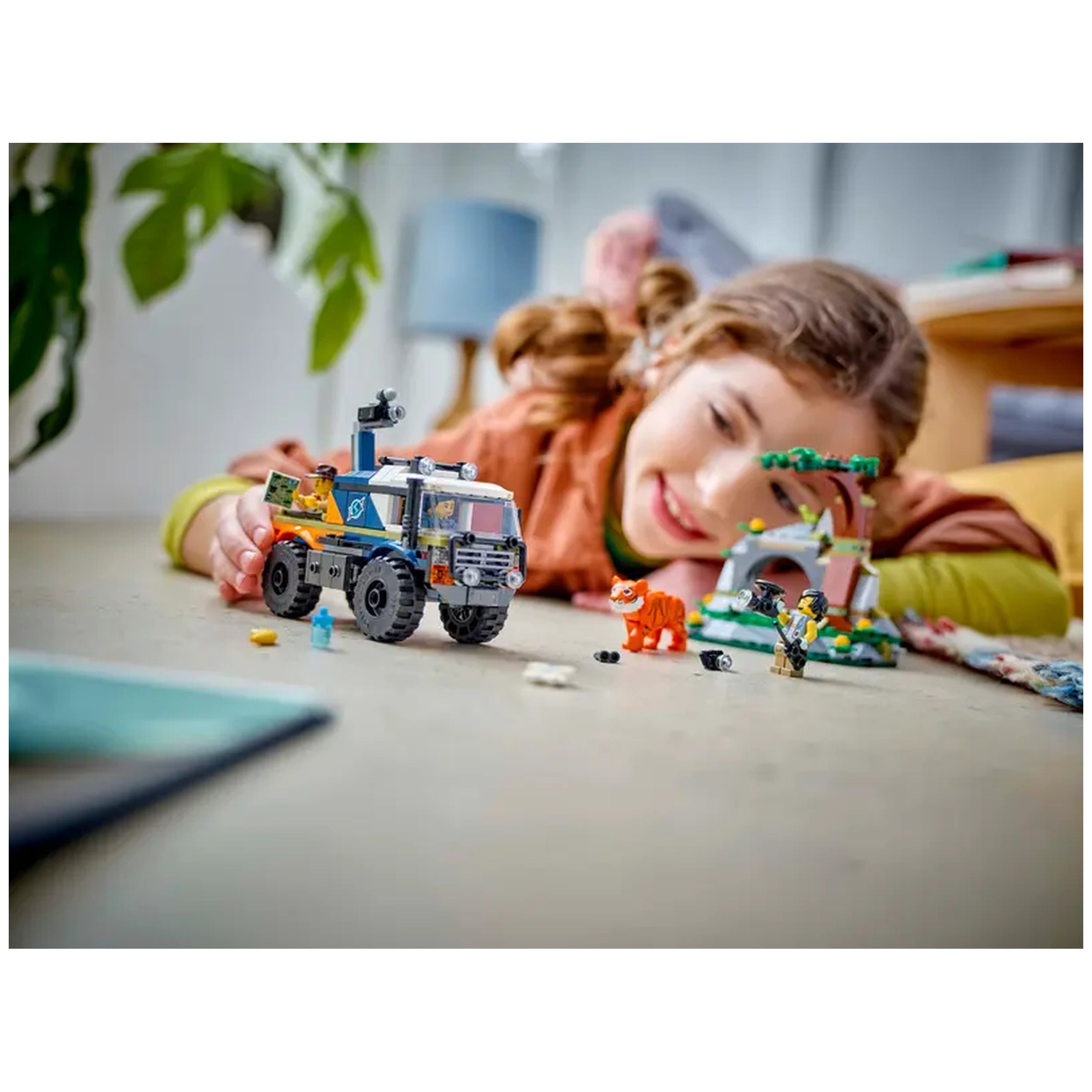 A young girl smiles while playing with the LEGO® City Jungle Explorer Off-Road Truck, surrounded by a vibrant jungle cave and explorer minifigures.