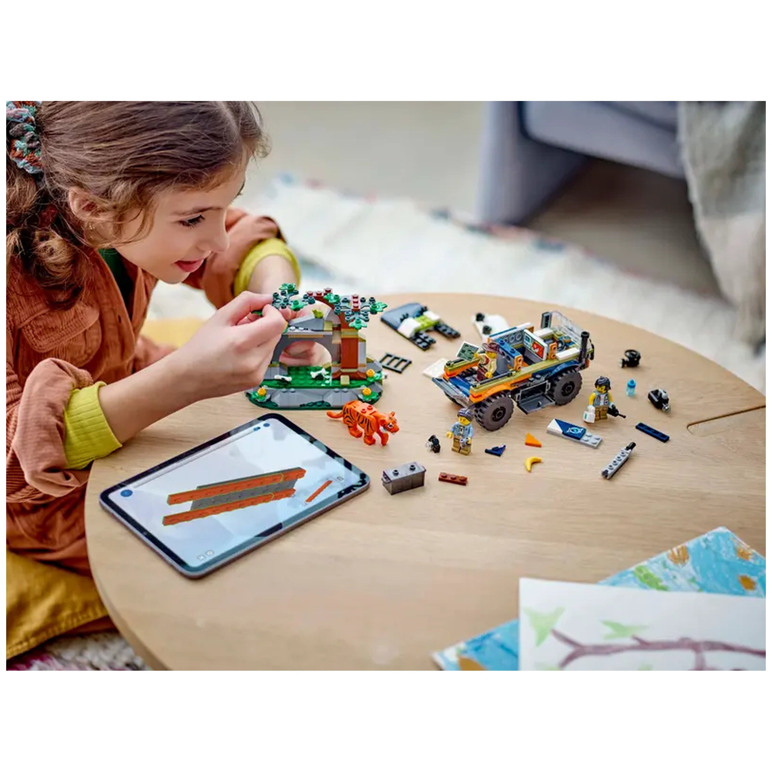 A child with wavy hair focuses on building a LEGO jungle scene on a wooden table, surrounded by colorful pieces, including an off-road truck, explorer minifigures, and a tiger figure. An interactive tablet with building instructions sits nearby, while a cozy living space is visible in the background.