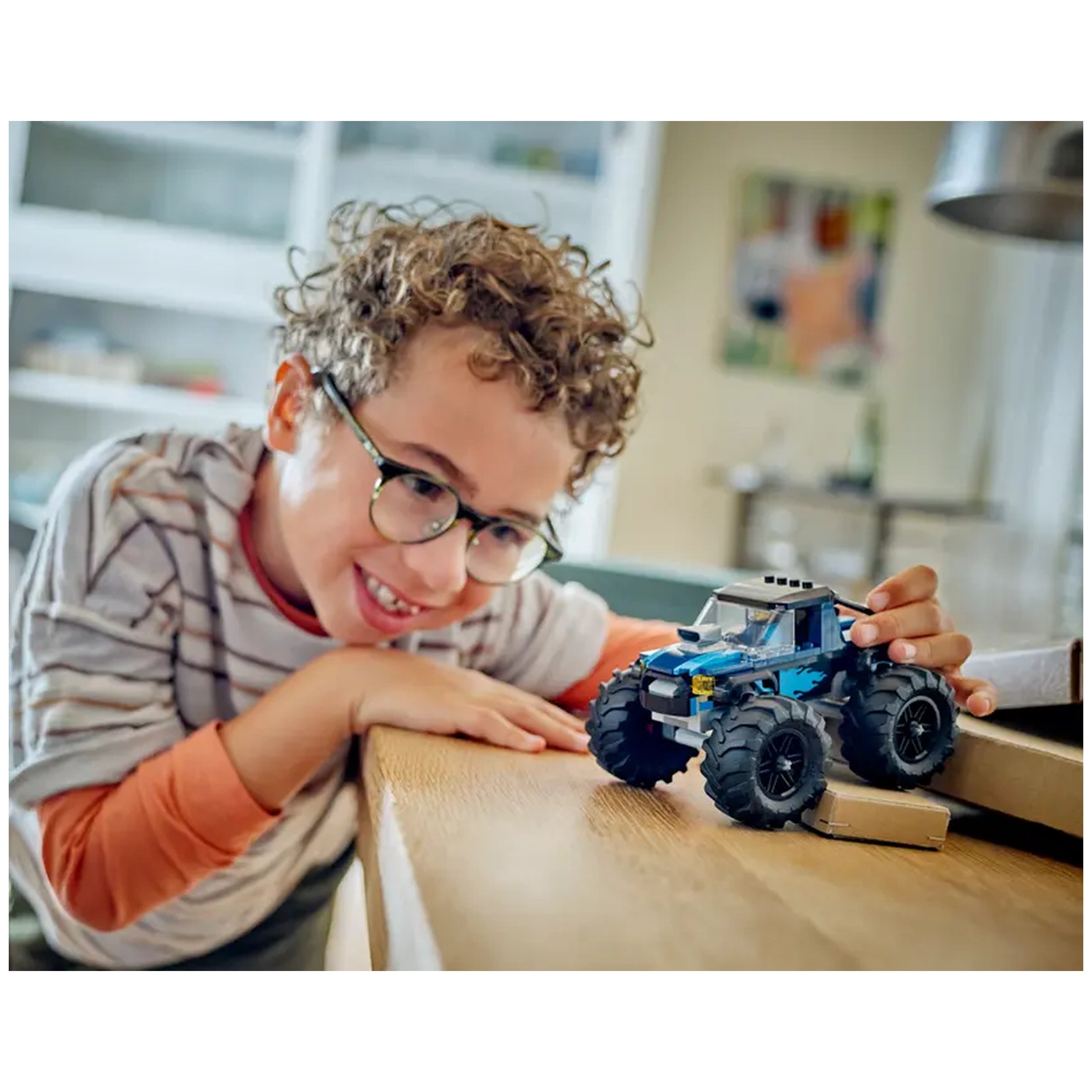 A smiling child with curly hair and glasses leans over a wooden table, excitedly playing with a blue LEGO City monster truck.
