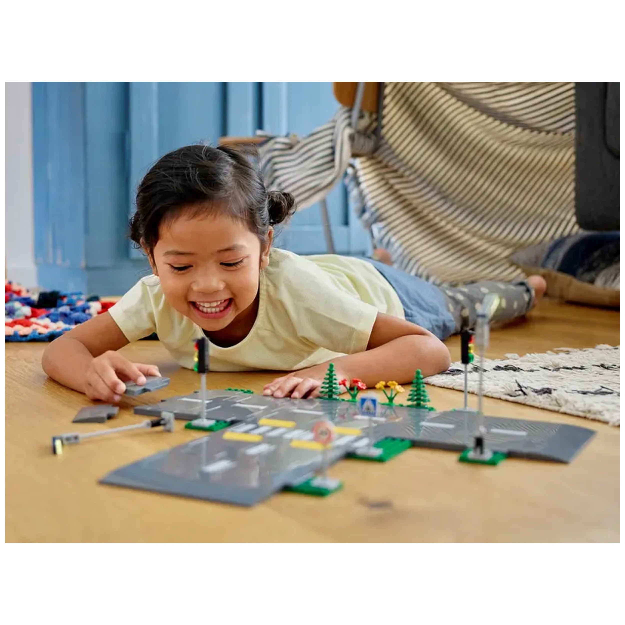 A young girl joyfully plays on the floor with LEGO City Road Plates, assembling road designs with trees, streetlights, and traffic signs.