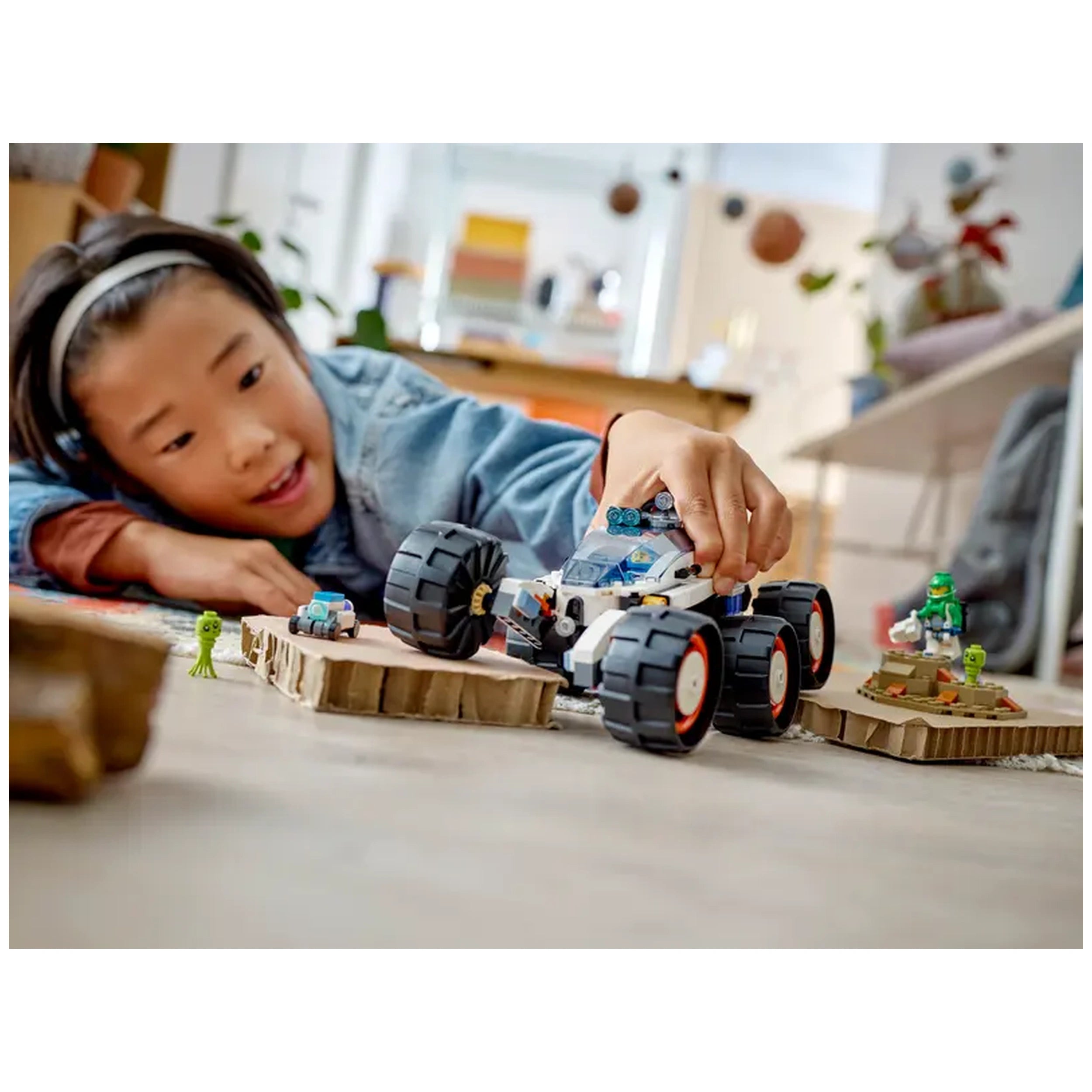 A child plays on the floor with the LEGO® City Space Explorer Rover, maneuvering it alongside alien figures on cardboard terrain.